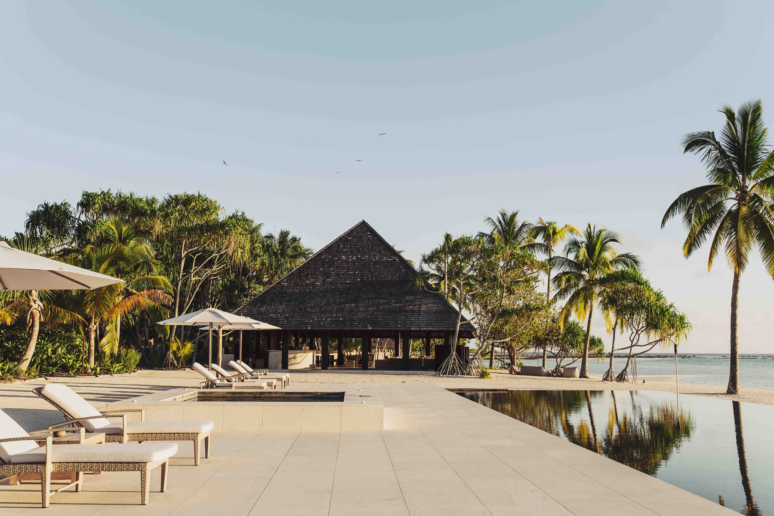 Plage tropicale avec chaises longues, parasols, piscine, cabane en bambou, palmiers et océan en arrière-plan.
