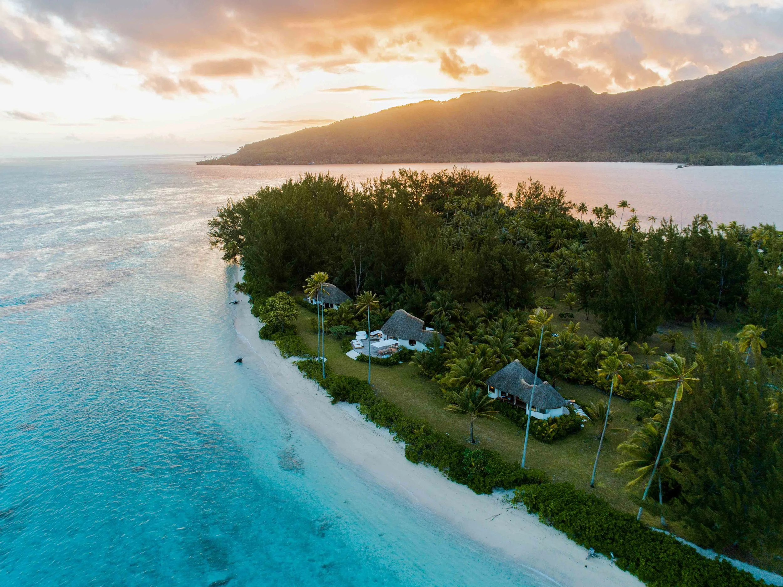 Vue aérienne d'une île tropicale avec des maisonnettes, entourée de mer turquoise, de palmier et de végétation dense au coucher du soleil.