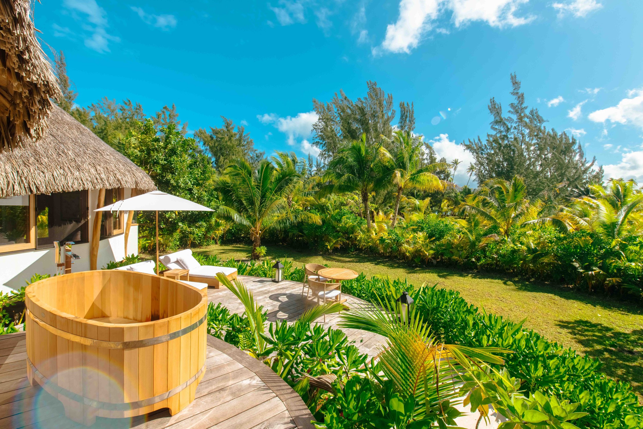 Terrasse en bois avec jacuzzi en bois, chaises longues avec coussins, parasol blanc, entourée de végétation tropicale dense sous un ciel bleu avec quelques nuages.