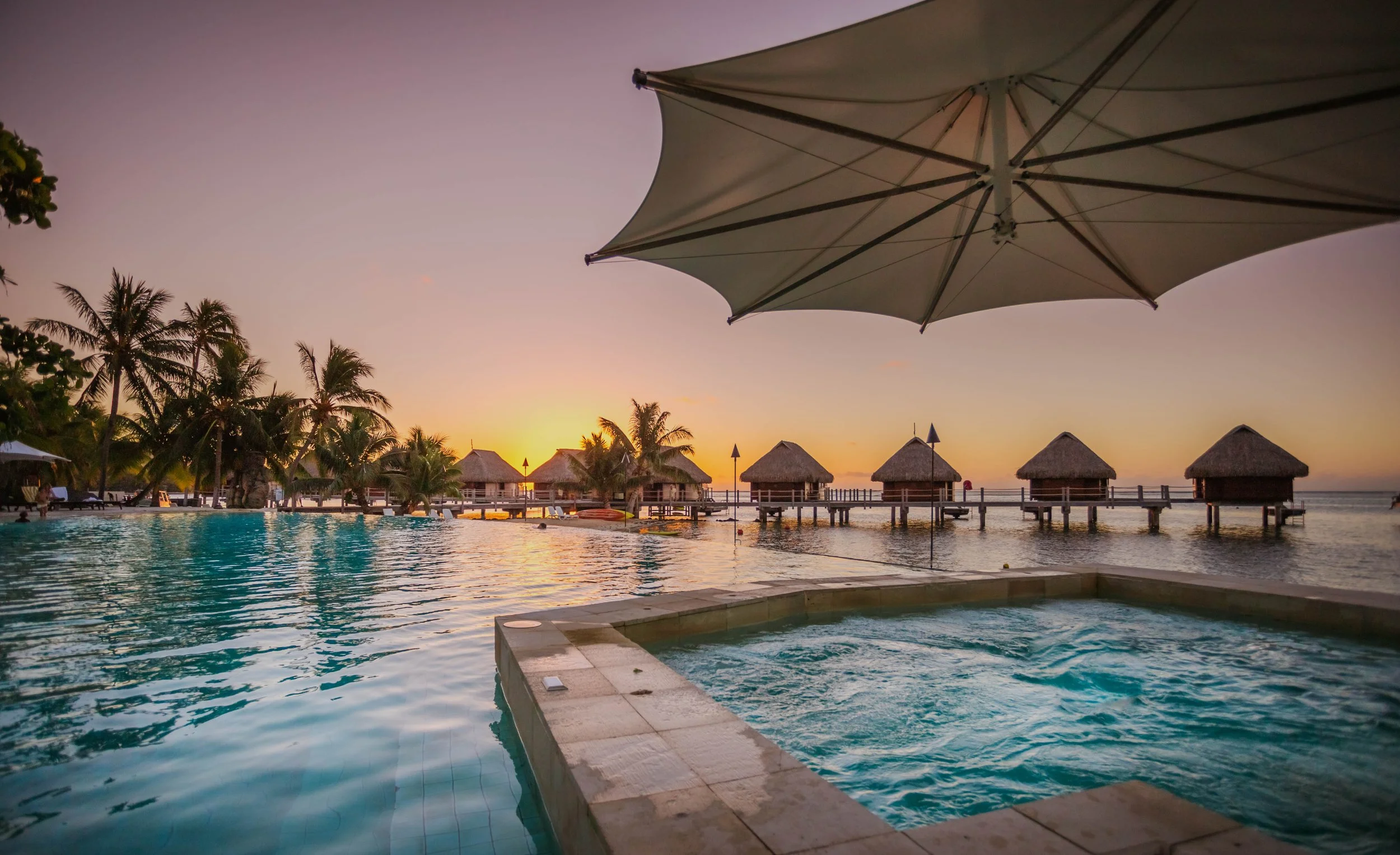 Piscine avec vue sur des cabanes sur pilotis en bord de mer au coucher du soleil, avec parasol sur la terrasse