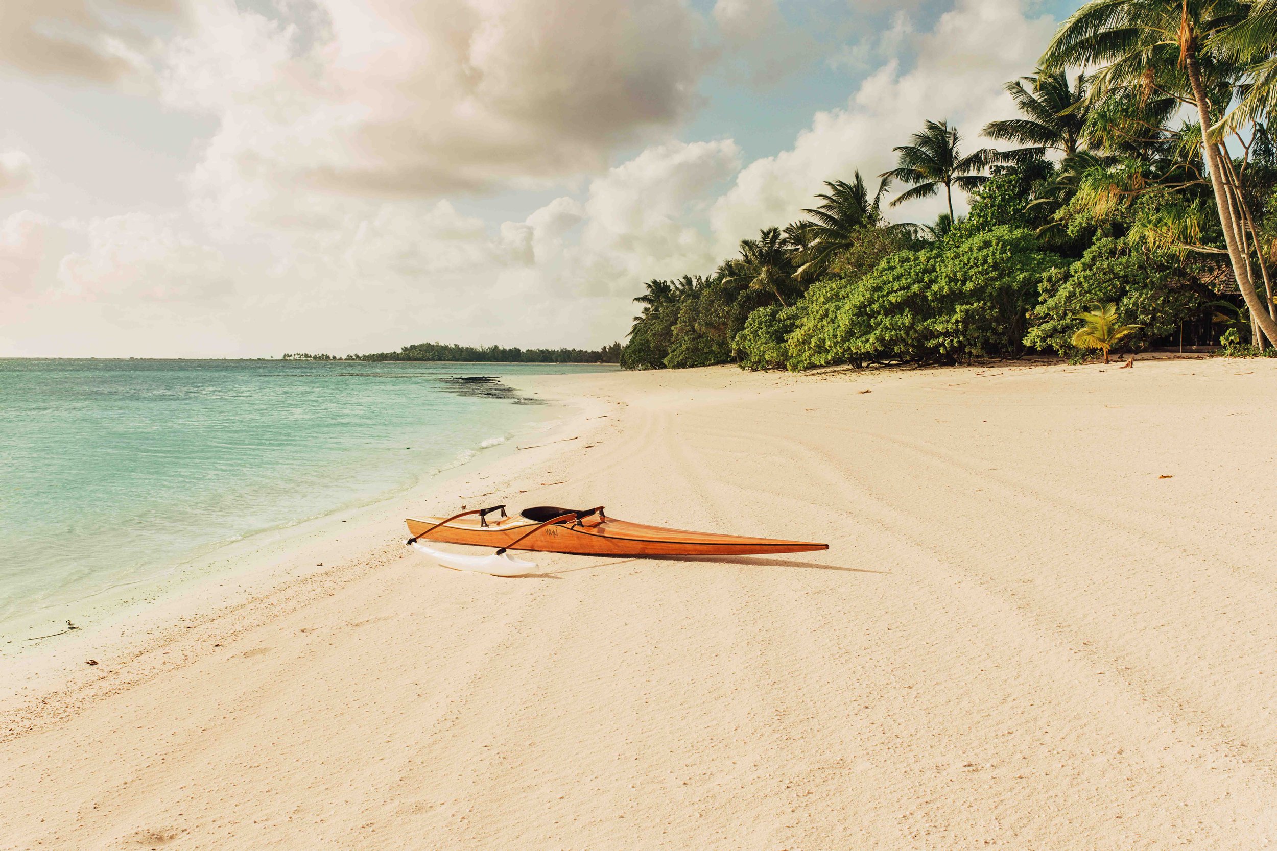 Une plage de sable blanc avec deux kayaks en bois abandonnés au bord de l'eau turquoise. À l'arrière, il y a une végétation dense de palmiers et d'arbres verts. Le ciel est partiellement nuageux.