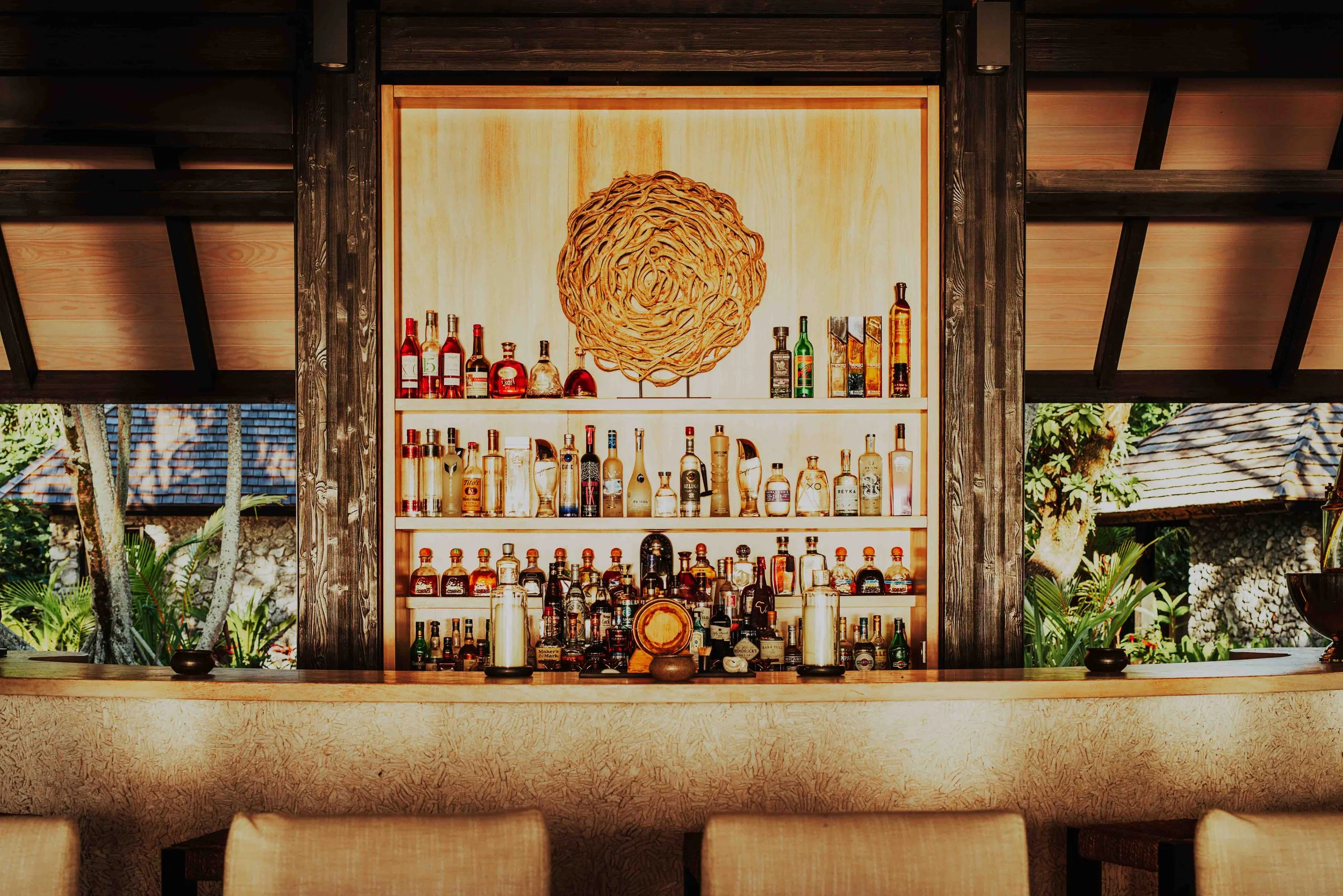 Bar intérieur avec étagère de bouteilles d'alcool, décor mural en bois et vue sur un jardin tropical.
