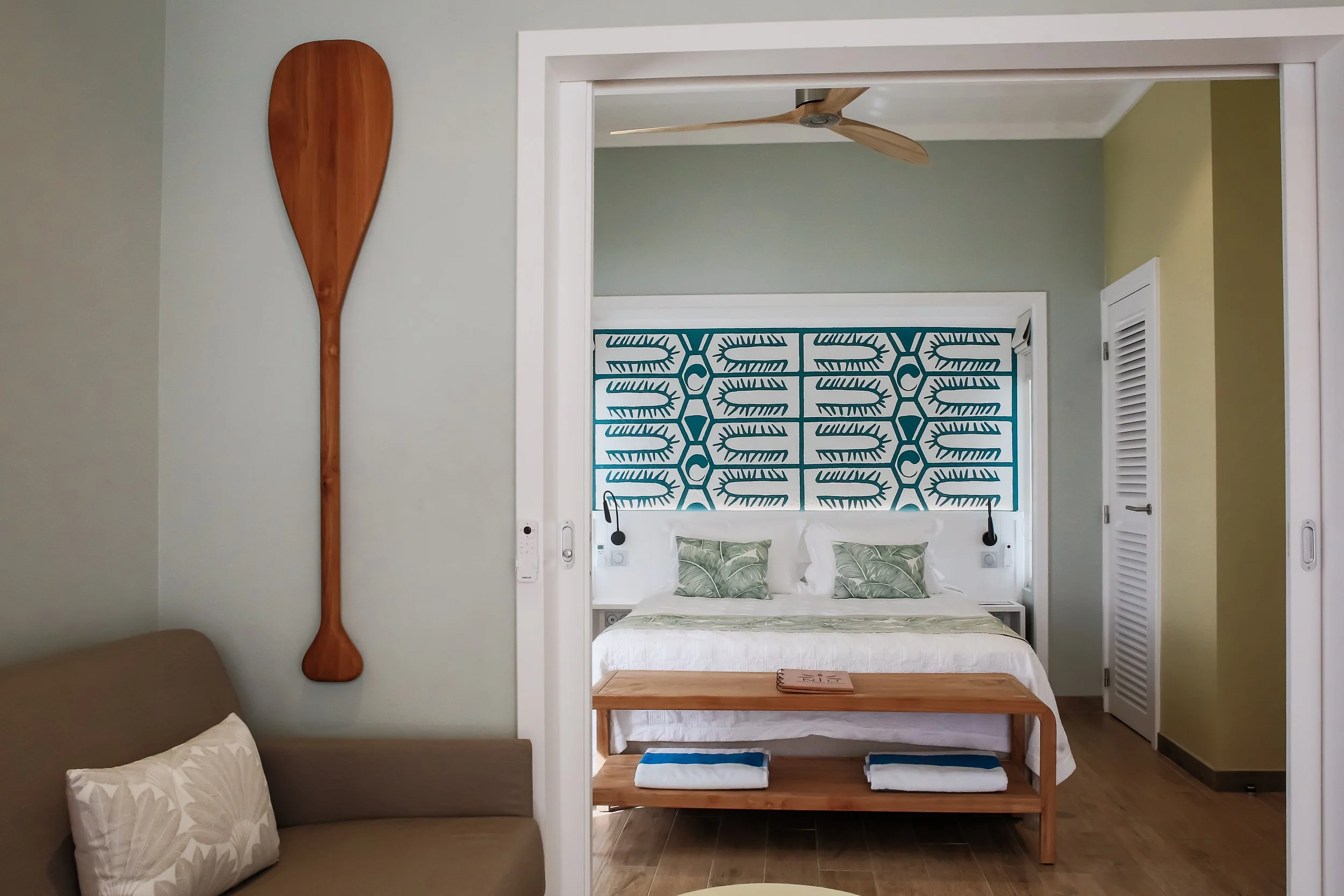 Chambre d'hôtel avec décoration tropicale, tête de lit bleue avec motif de feuilles, lit avec linge blanc et coussins à motifs de feuilles, meuble en bois avec serviettes, mur avec décoration en bois en forme de paddle, plafond avec ventilateur en bo