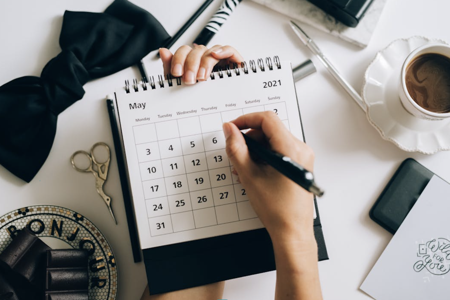 Person writing on a May 2021 calendar with a black pen, surrounded by office supplies, a cup of coffee, scissors, and a smartphone on a white desk.