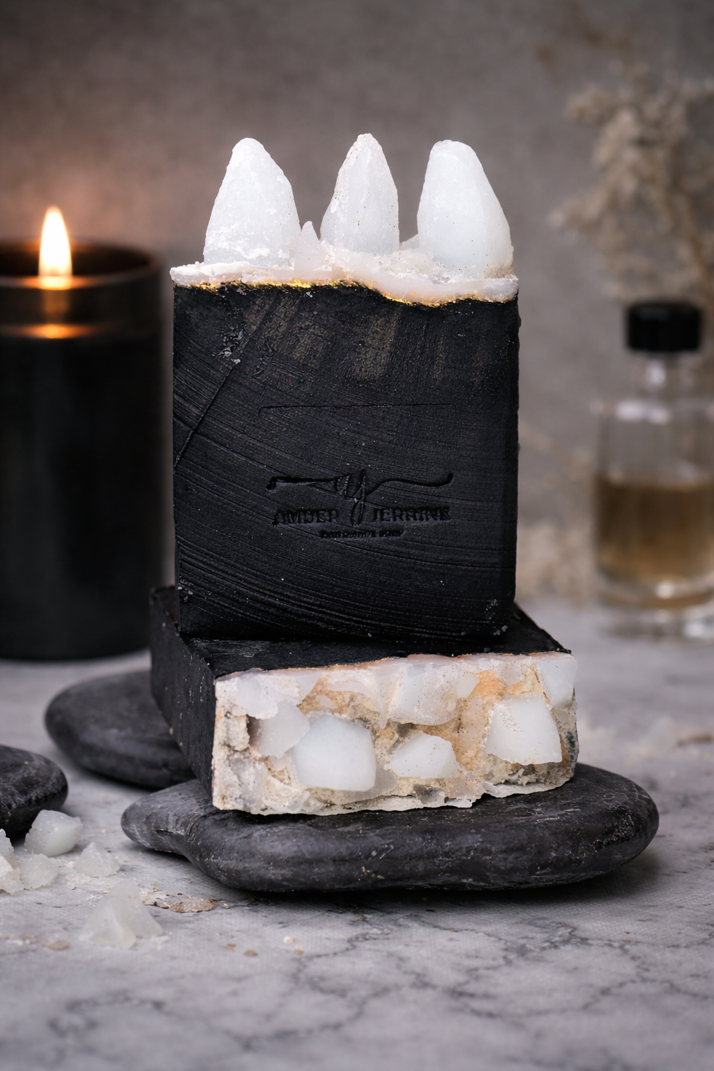 Handcrafted black soap bar with white crystal like embeds on top, accented with gold mica, styled on smooth stones with a candle in the background.