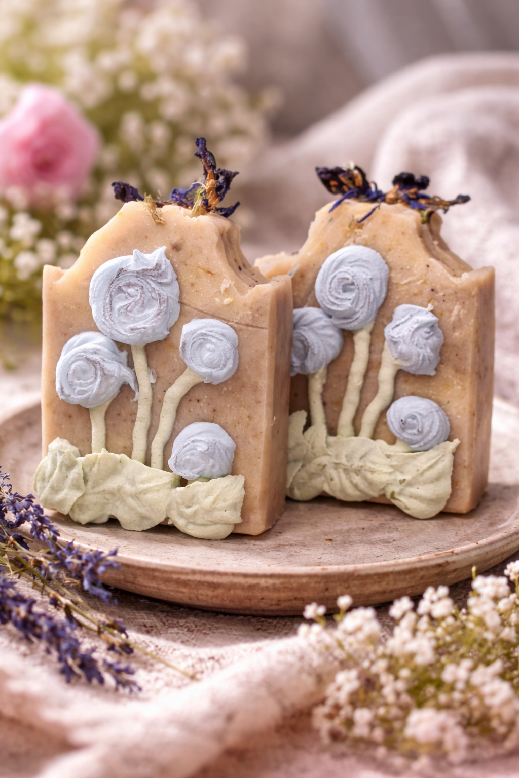 Two Second Bloom soap bars on ceramic plate with blue piped roses, green leaves, and dried botanicals on warm beige base. Lavender and white flowers in soft garden setting.