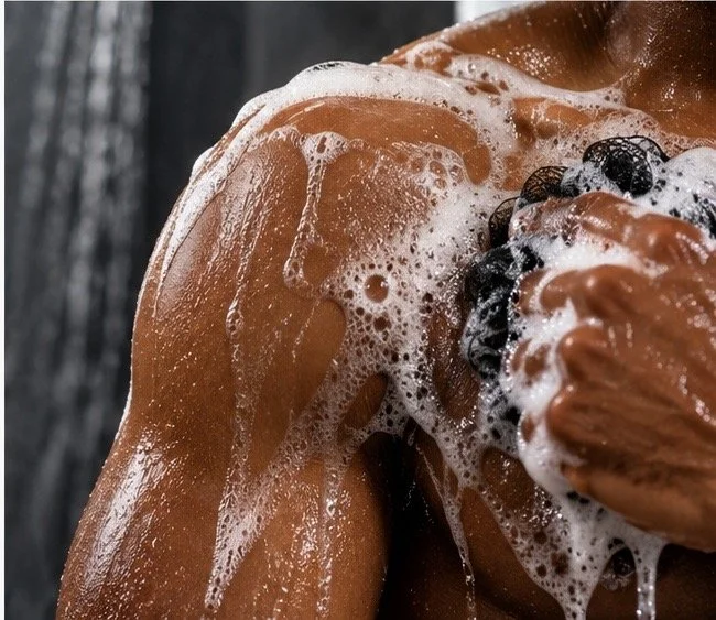 Person cleansing their shoulder with rich body wash lather and a black shower pouf, highlighting the creamy foaming experience of Amber Jerrine Mr. Right Luxury Body Wash.