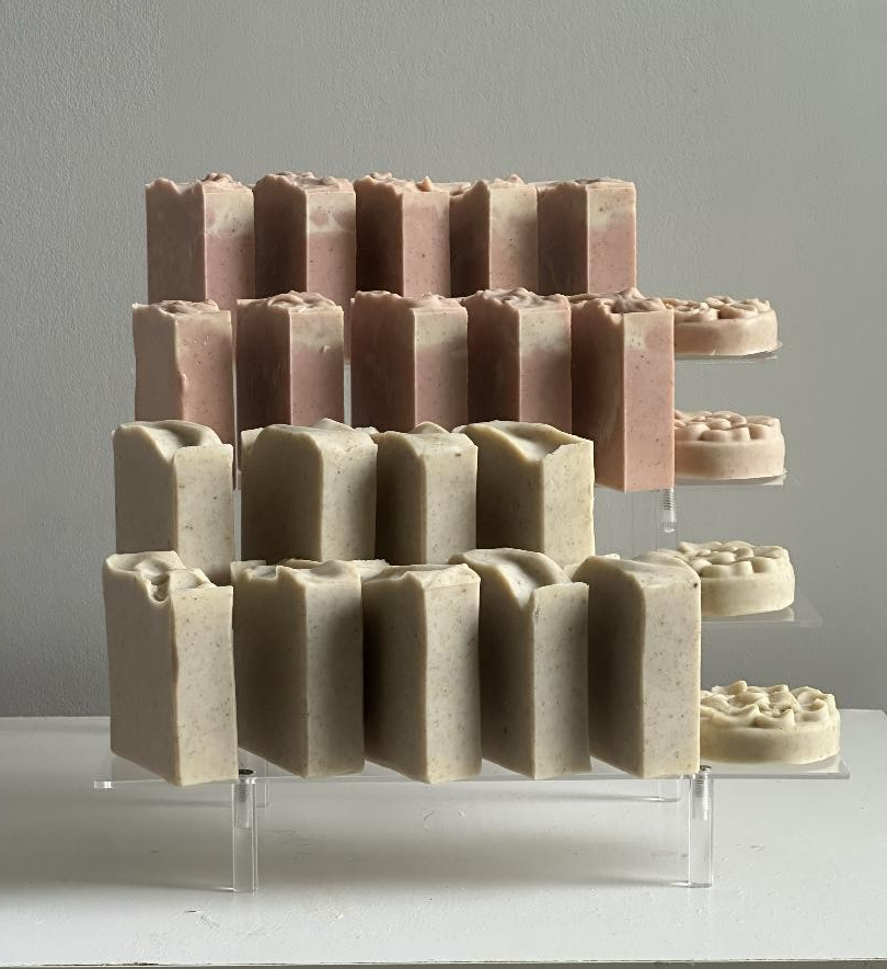 Three-tier display of various handcrafted soap bars in shades of pink, beige, and white, arranged on a clear acrylic stand.