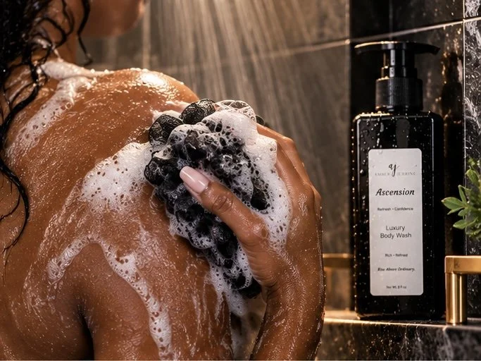 Woman cleansing her shoulder with rich foamy lather and a black shower pouf, Ascension Luxury Body Wash displayed beside her in an elegant shower setting.