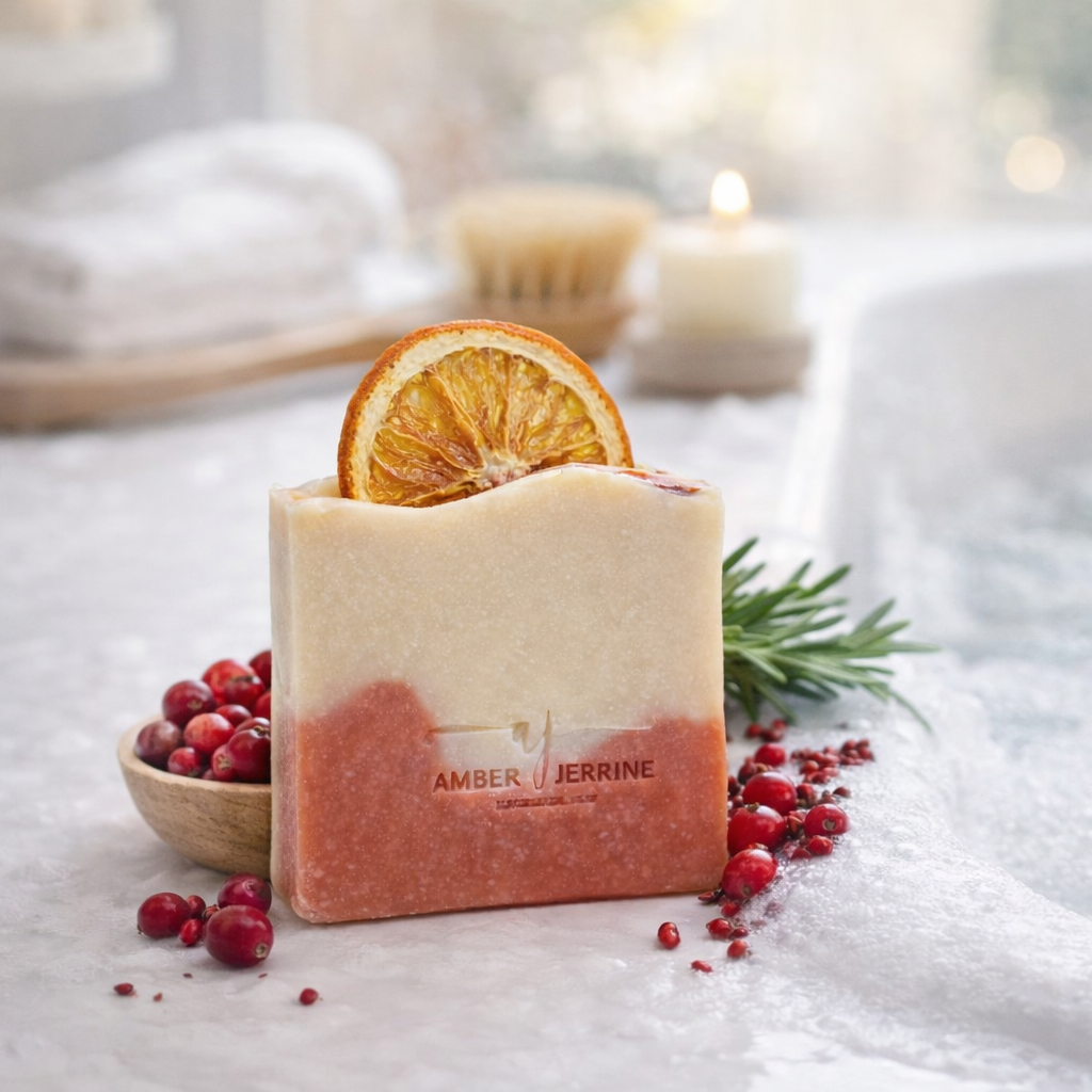 Two-tone handmade soap bar in cream and cranberry hues topped with a dried orange slice, styled beside red berries and greenery near a bubble-filled bathtub with a candle and towels in the background.