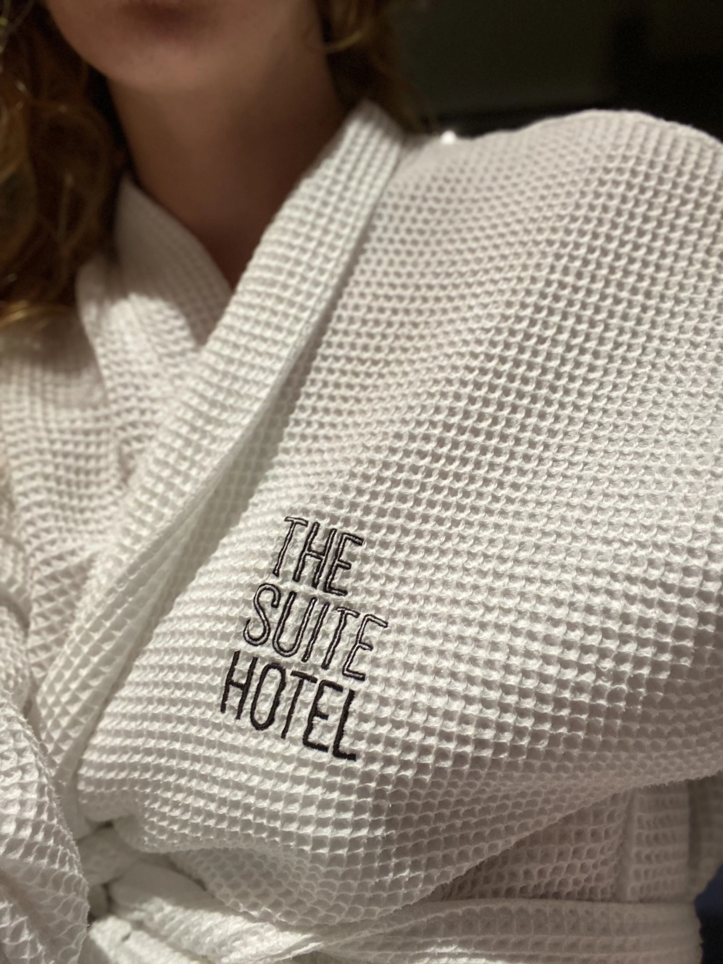 Nahaufnahme eines weißen Bademantels mit der Aufschrift 'The Suite Hotel' auf der linken Brustseite.