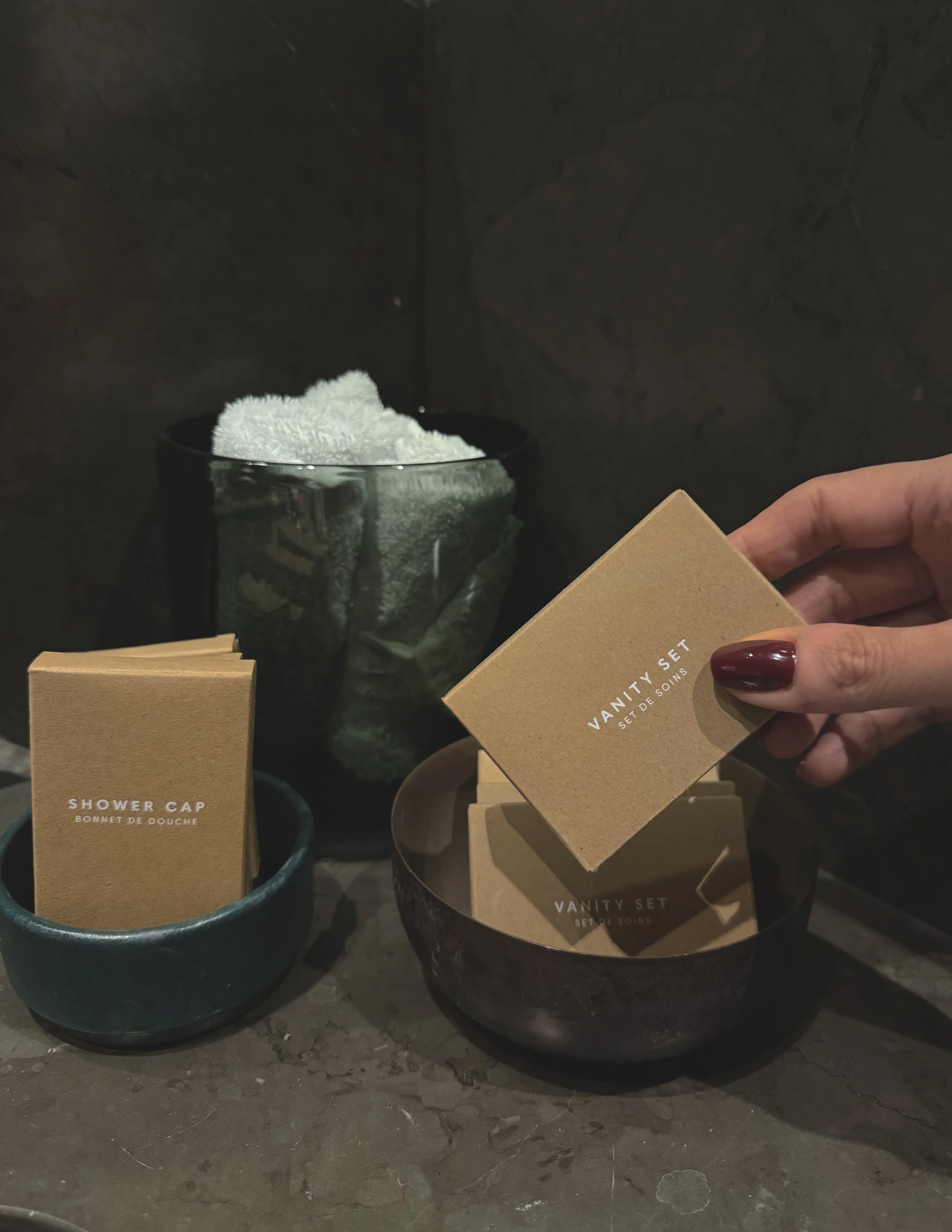 Hand hält eine kleine Schachtel mit dem Etikett "VANITY SET", neben einer Schale mit weiteren "VANITY SET" Packungen, vor einem dunklen Hintergrund mit Steineffekt.