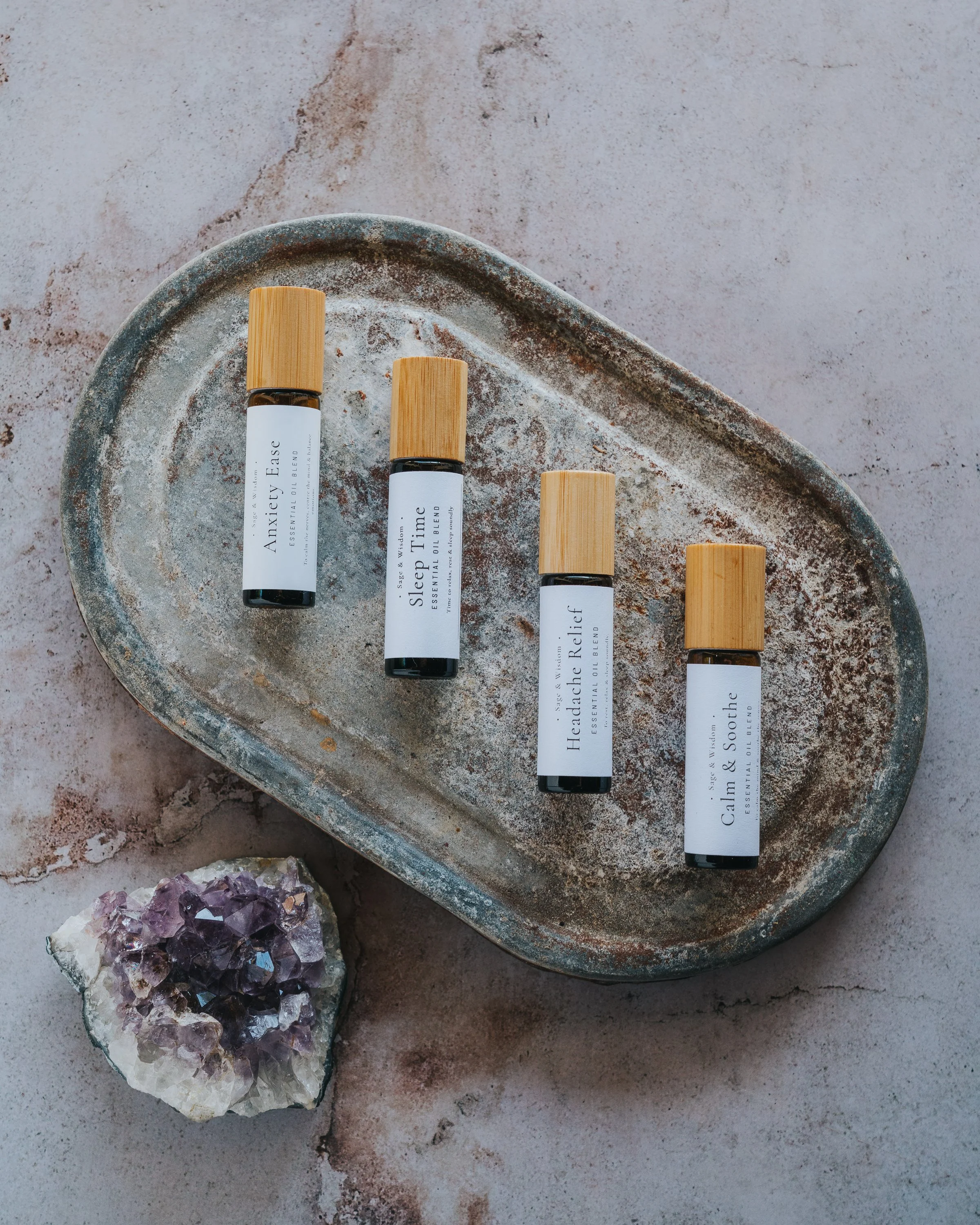 Four essential oil bottles with wooden caps on a textured oval tray, next to a crystal cluster on a pinkish surface.