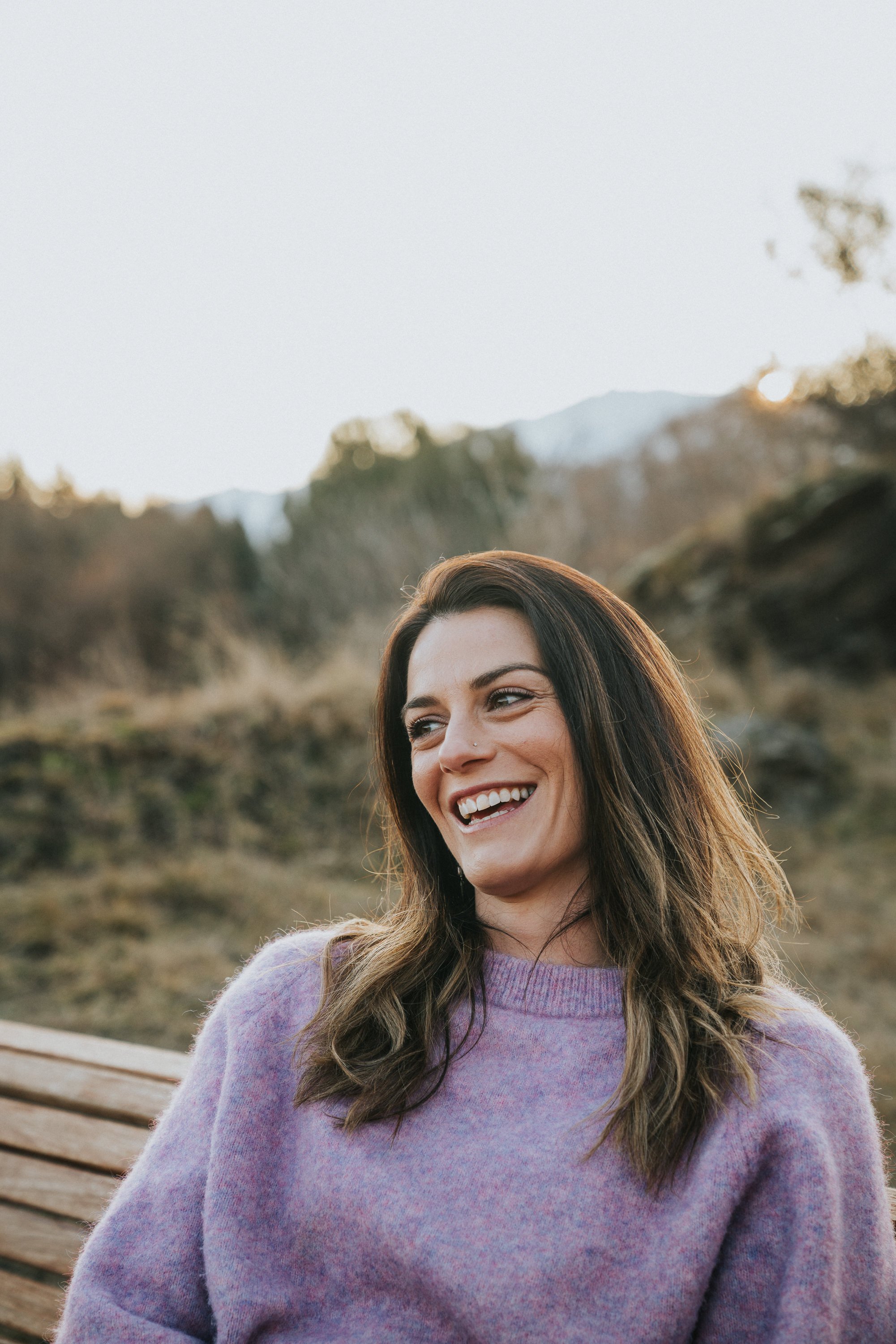 A woman with dark hair in loose waves, wearing a lavender sweater, smiling outdoors on a bench with mountains and trees in the background during the daytime.