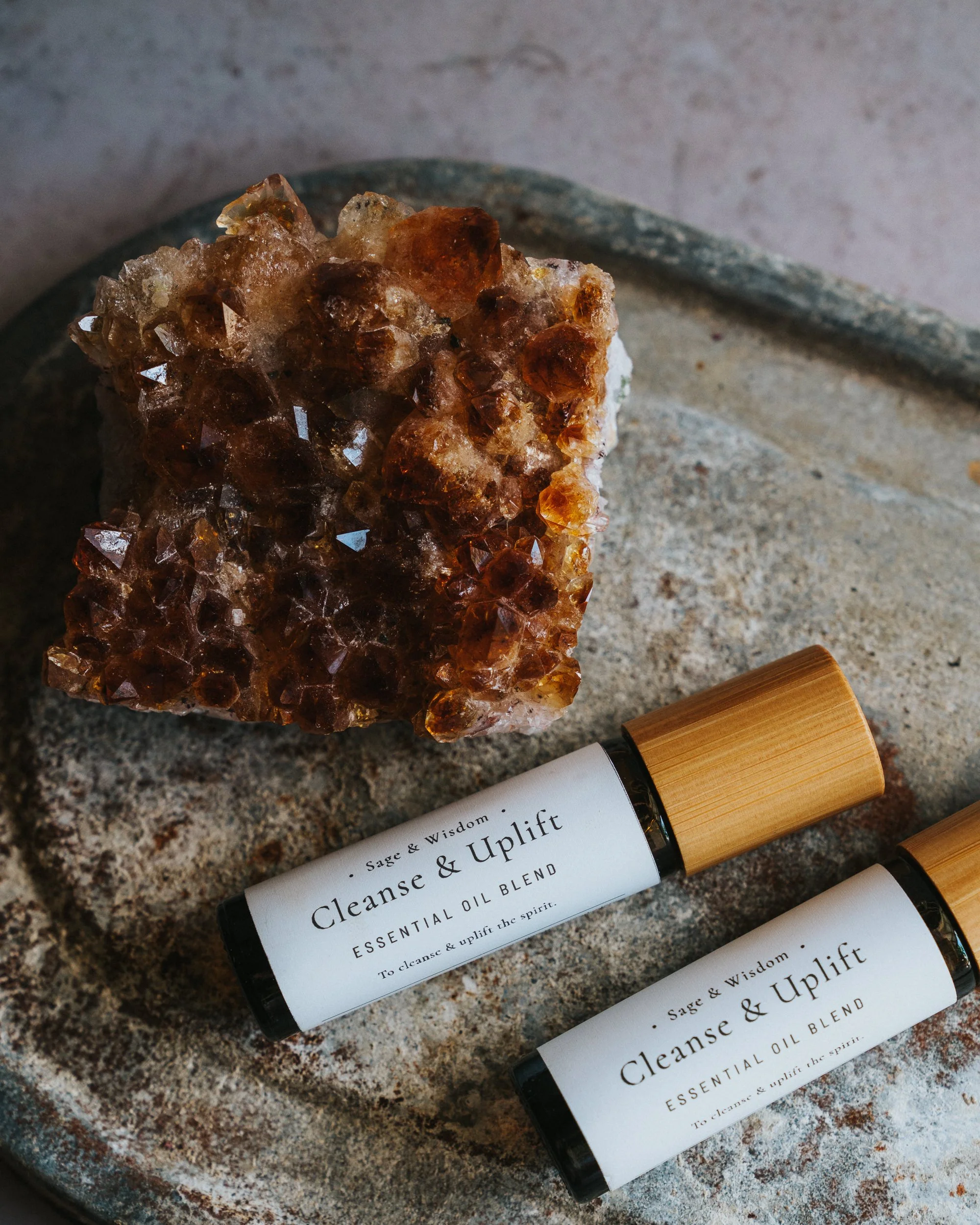 A large orange-brown crystal cluster next to two small bottles of essential oil labeled 'Cleanse & Uplift' on a round textured gray surface.