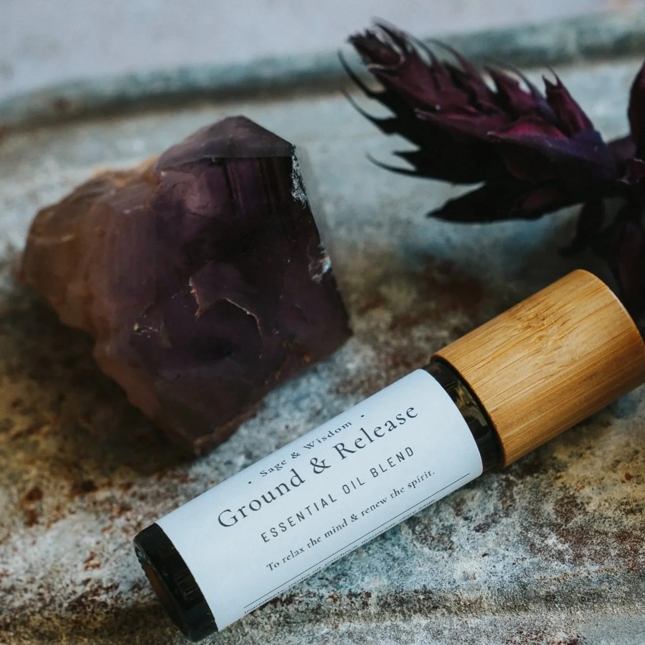 A bottle of Ground & Release essential oil blend lying on a rugged surface, accompanied by a purple flower and a raw mineral or crystal.