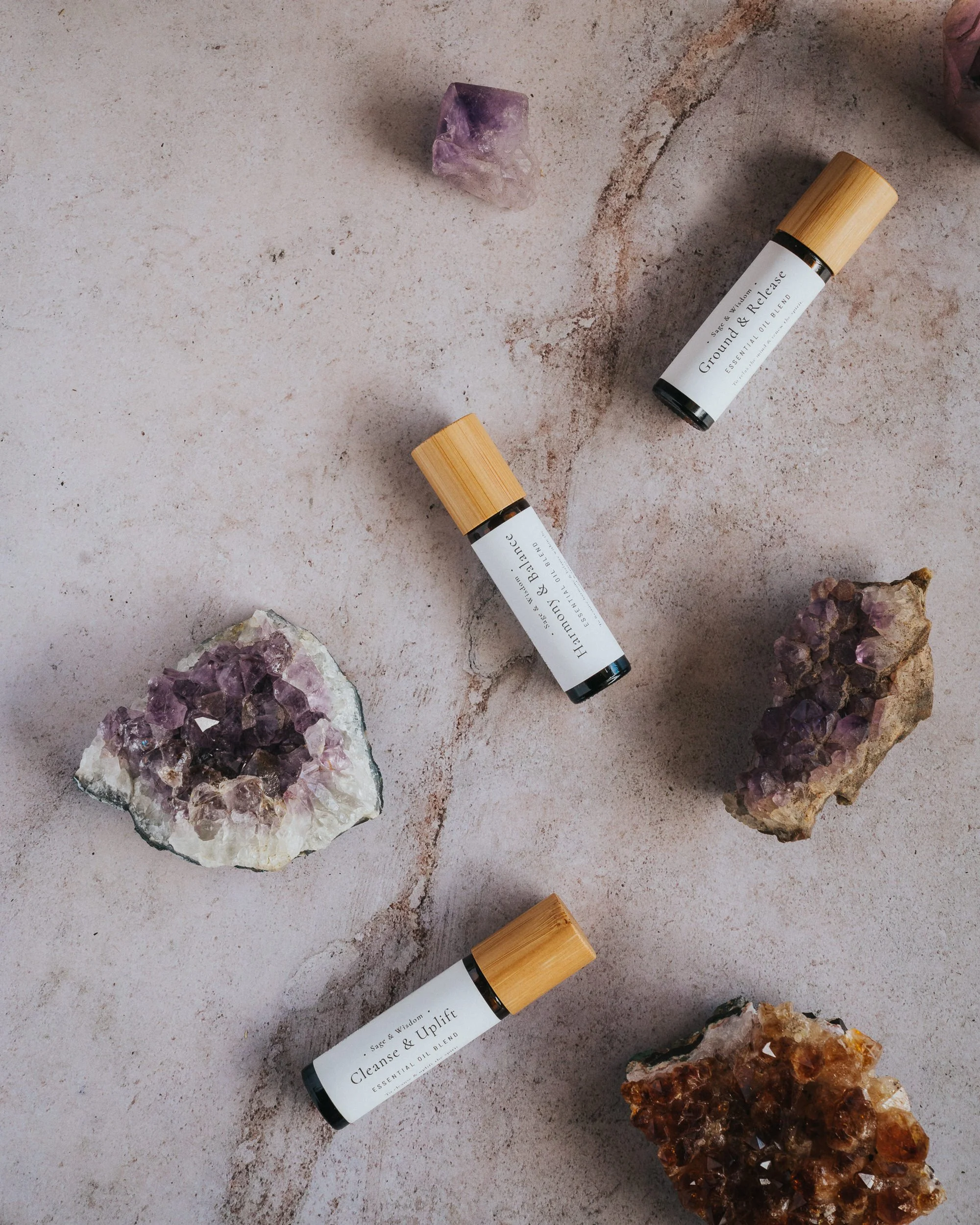 Three essential oil bottles with bamboo caps and white labels surrounded by amethyst crystals on a textured surface.