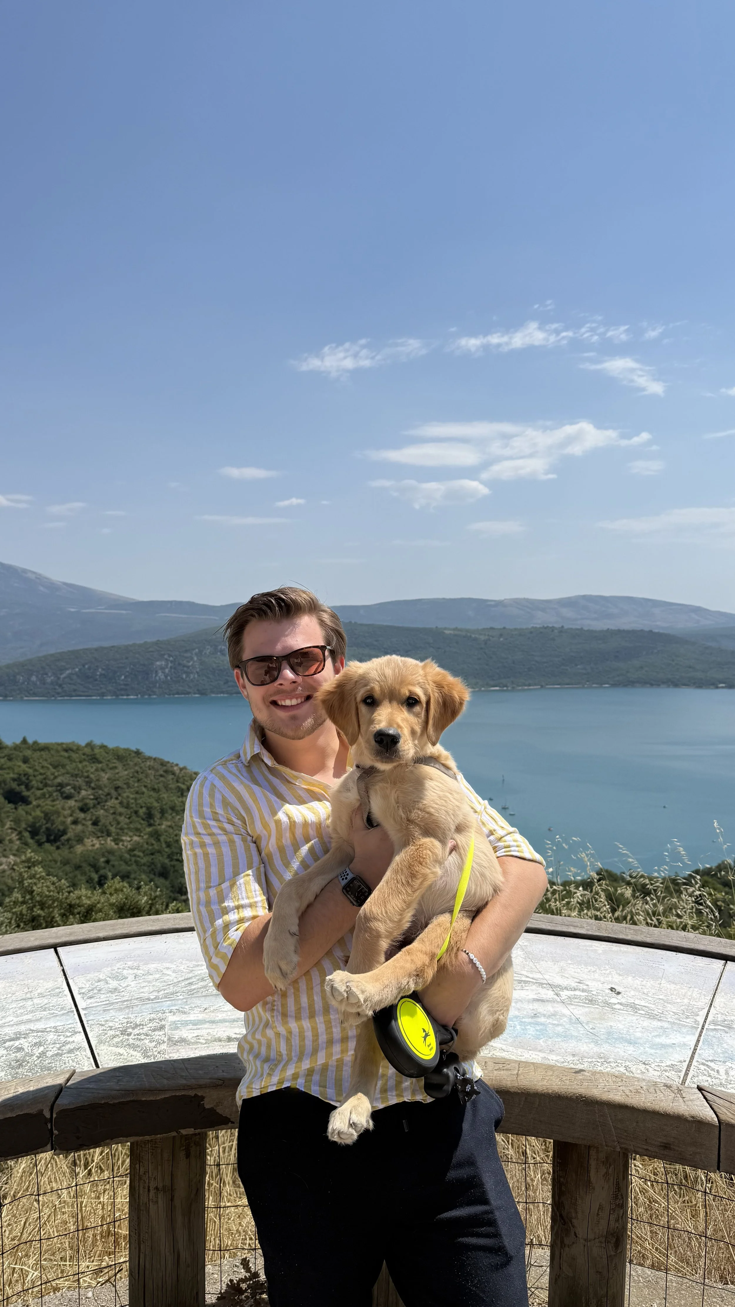Man met een Labrador puppy op een uitzichtpunt met een meer en bergen op de achtergrond