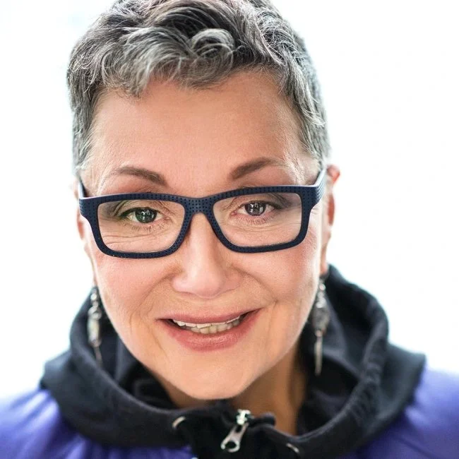 Close-up of a smiling woman with short gray hair wearing glasses and a dark jacket.