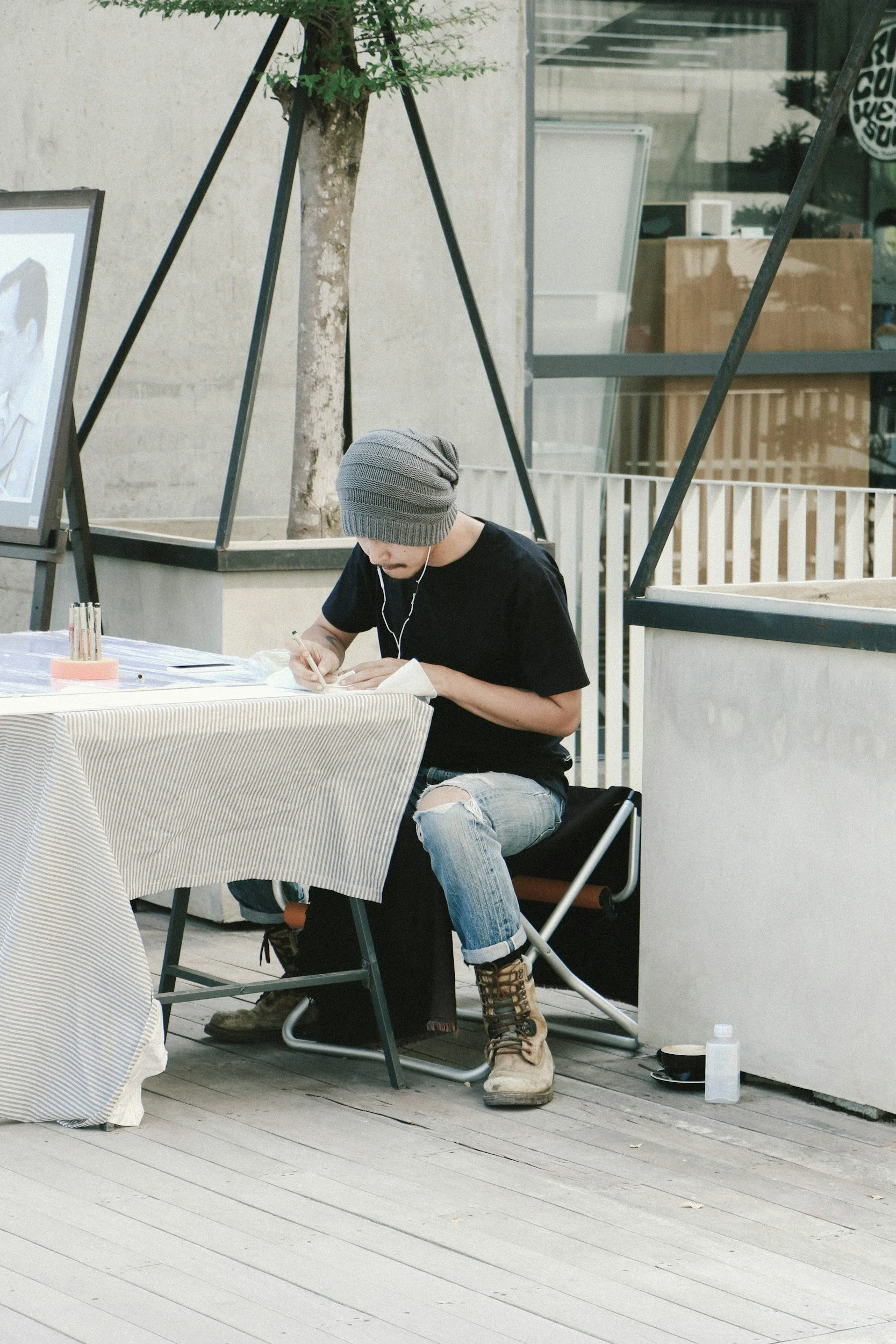 A person with a gray beanie, black T-shirt, ripped jeans, and tan boots sitting on a portable folding chair, writing at a table outside a modern building.