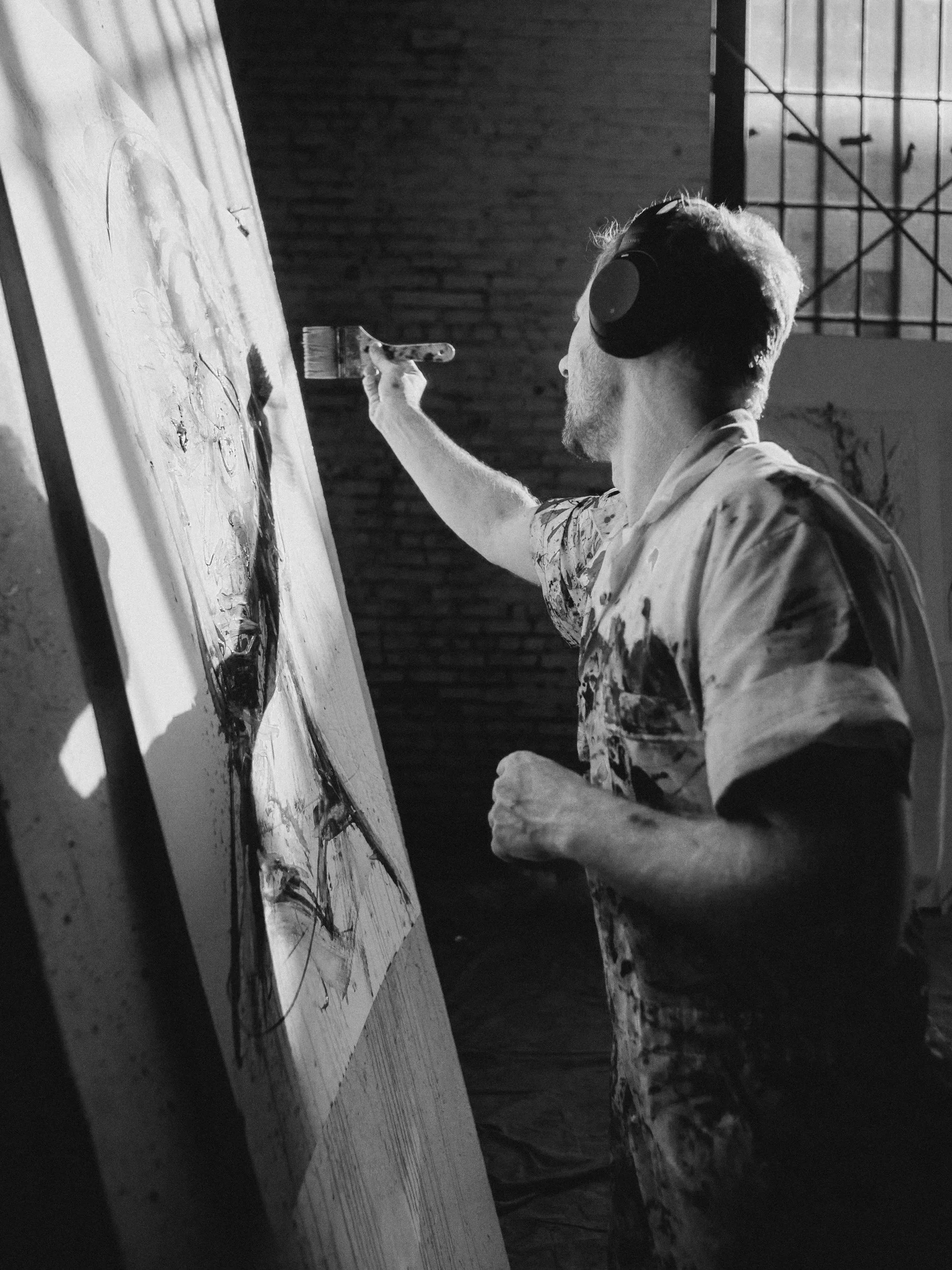 A man wearing headphones and a paint-splattered shirt is painting on a canvas, viewed from the side in a dimly lit studio with brick walls.