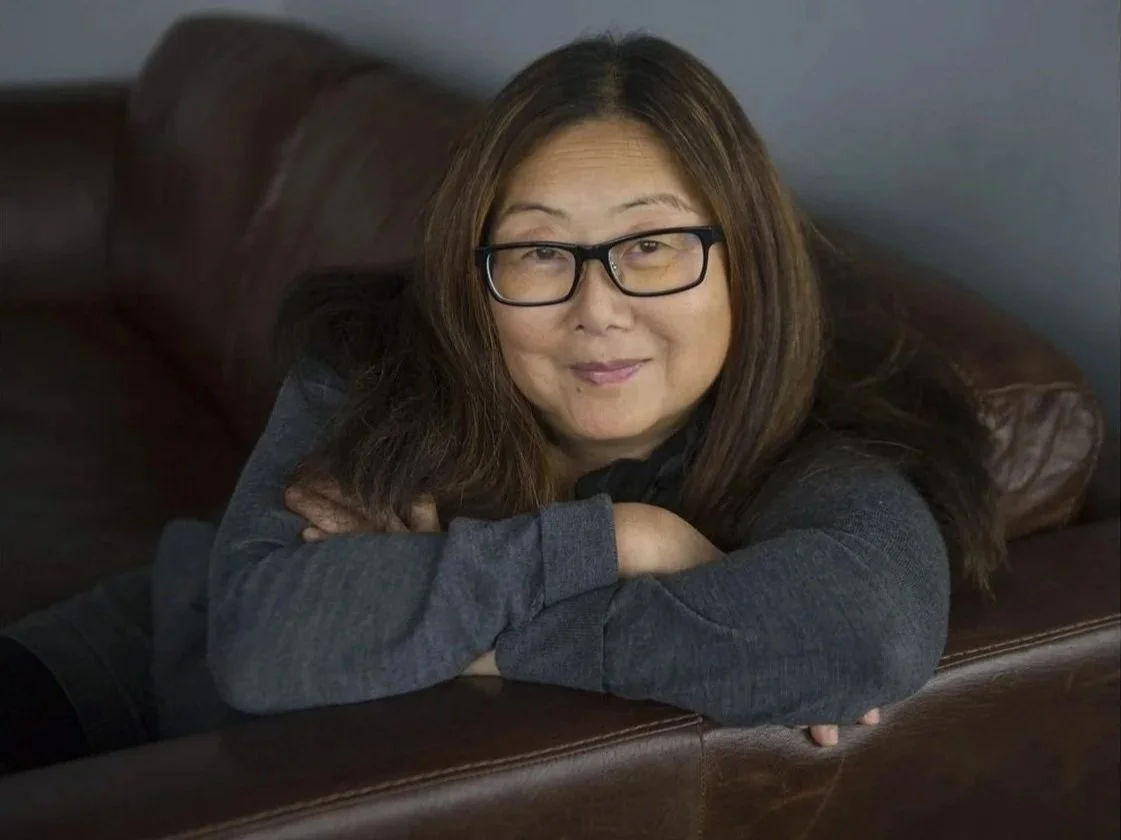 A woman with long brown hair, glasses, and a gray sweater leaning on a leather couch with her arms crossed, smiling at the camera.
