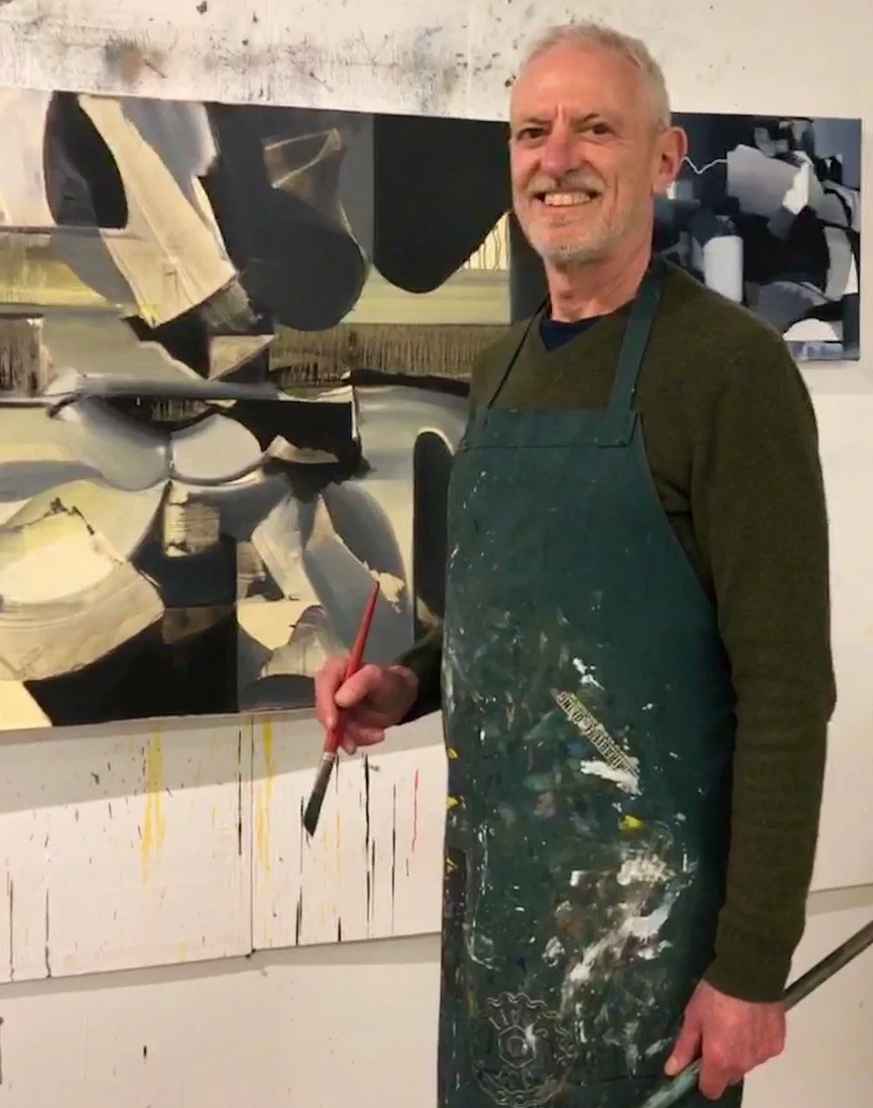 An elderly man with gray hair and a beard smiling, wearing a dark green shirt and a paint-splattered apron, stands in front of an abstract painting. He holds a paintbrush in his right hand.