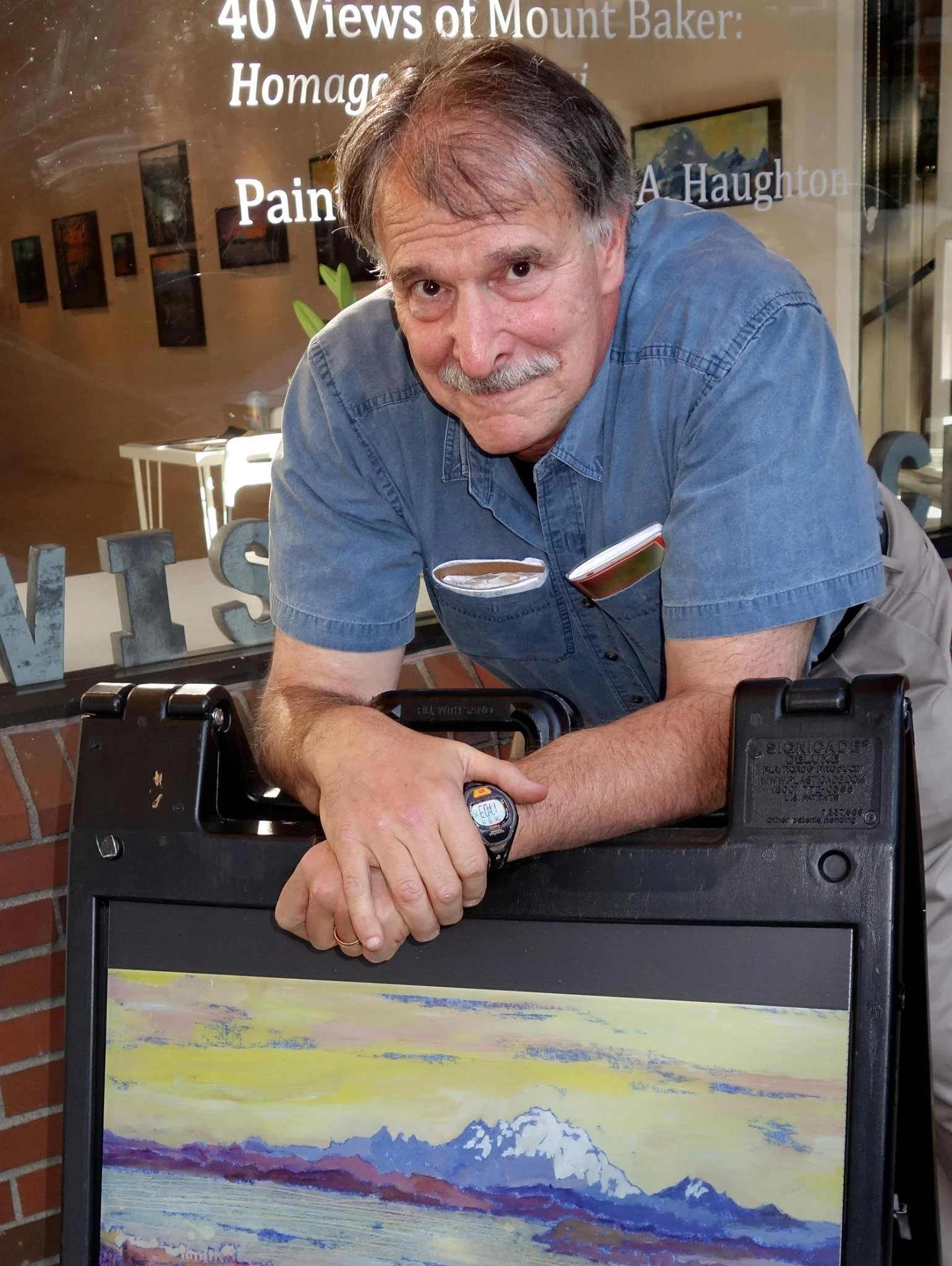 A man with gray hair and a mustache, wearing a blue short-sleeve shirt, leaning on a black sandwich board outside a building with a brick wall. The board displays a landscape painting of mountains and a lake. In the background, there are frames on the wall inside the building and a glass window with white text about Mount Baker.