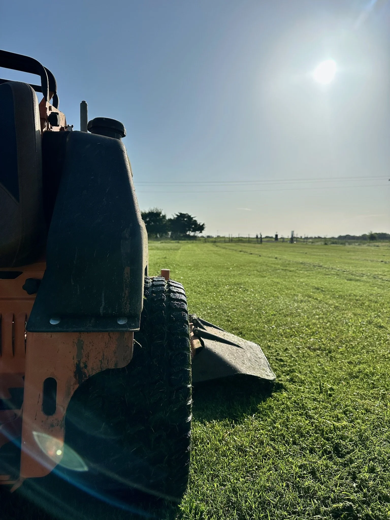 Close-up of a lawn mower on a freshly cut grassy field with trees in the background, under bright sunlight in the sky.