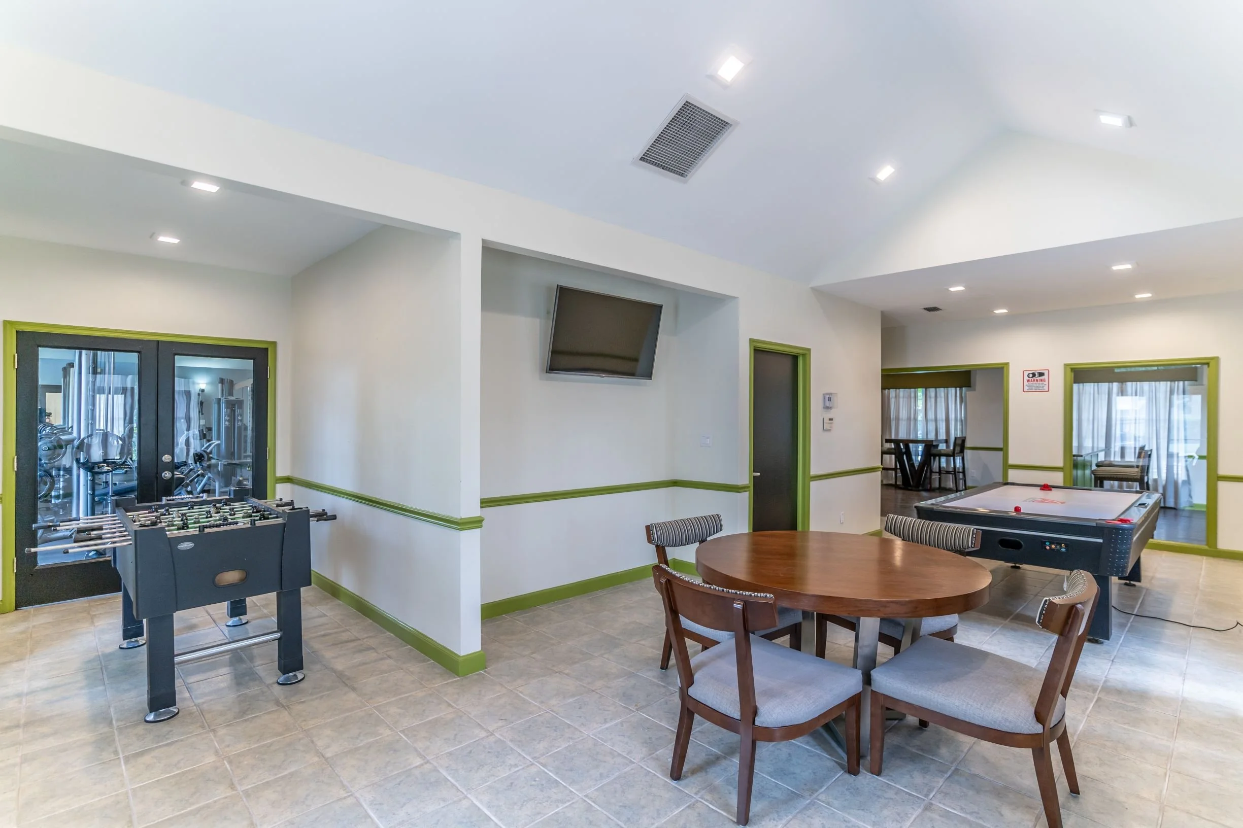 A recreational room with a ping pong table, a foosball table, a table with three chairs, and a television mounted on the wall, with large windows and glass doors.