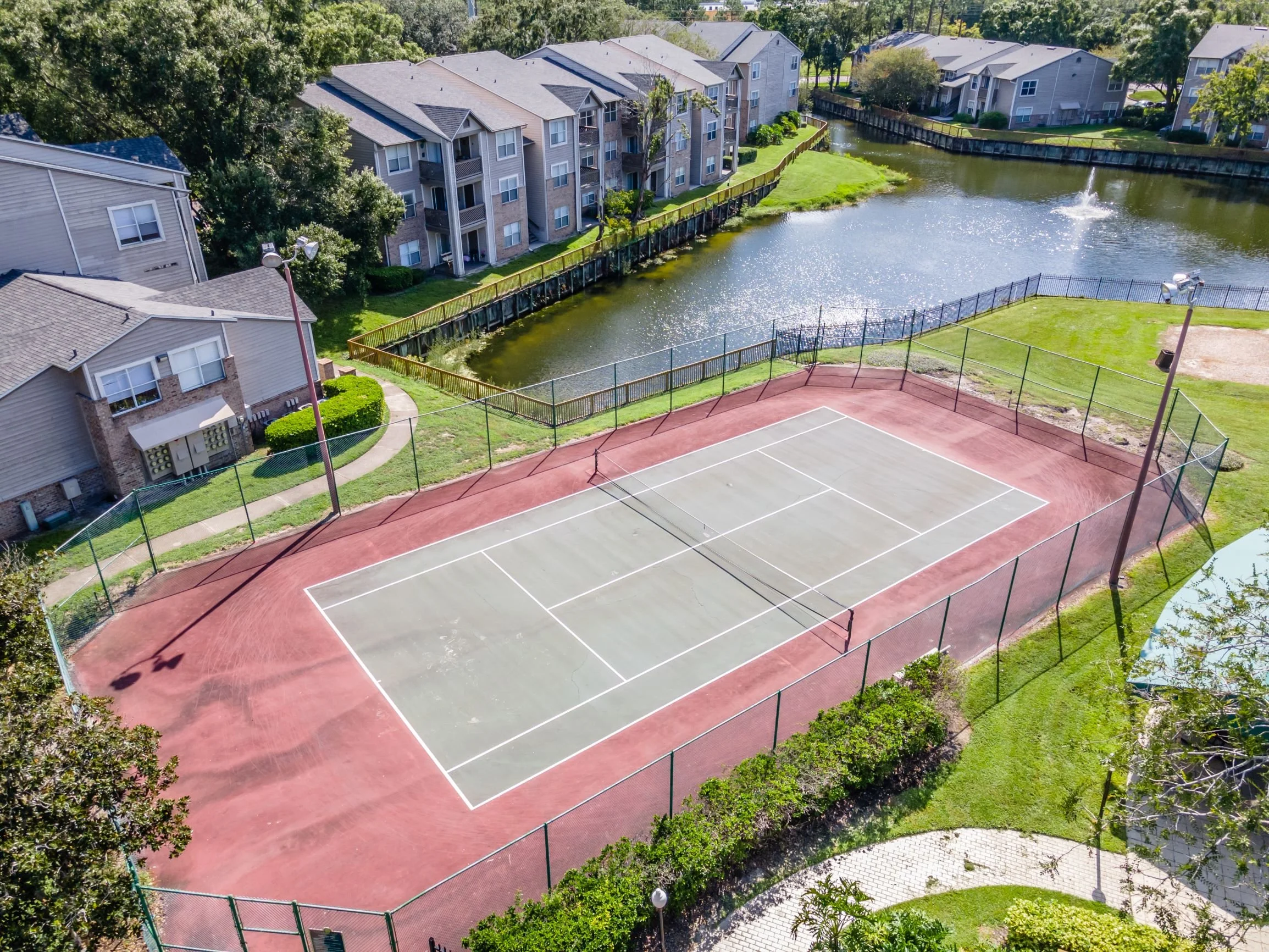 An outdoor tennis court surrounded by a black fence, adjacent to a small pond with a fountain, in a residential complex with multi-story apartment buildings and green lawns.