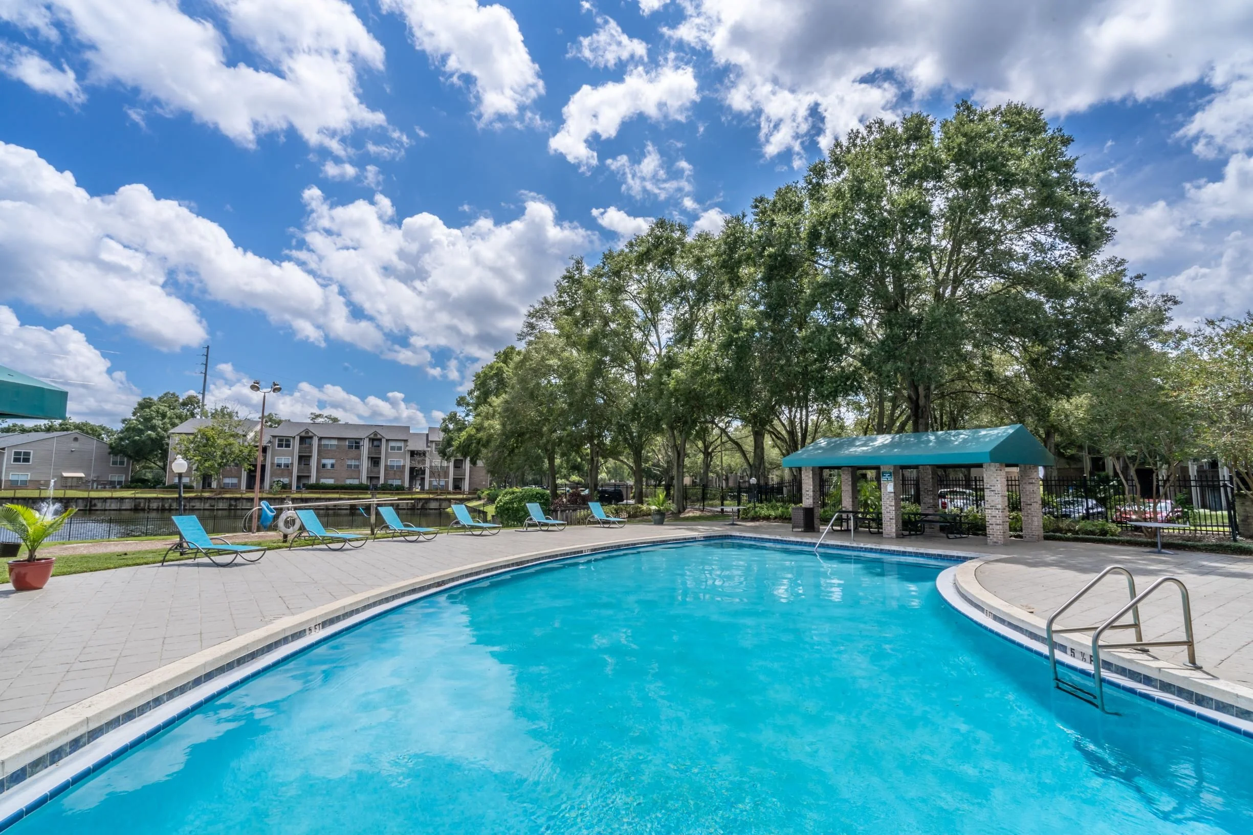 Outdoor swimming pool with blue lounge chairs, a shaded picnic area, and trees under a partly cloudy sky.