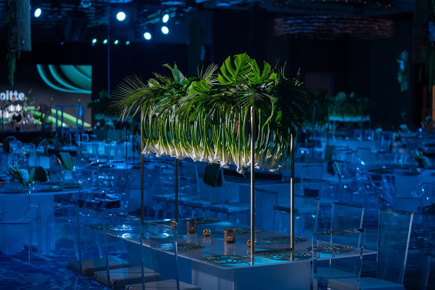 Indoor event space with tables, chairs, and a large floral centerpiece of green tropical leaves and white flowers under blue lighting.