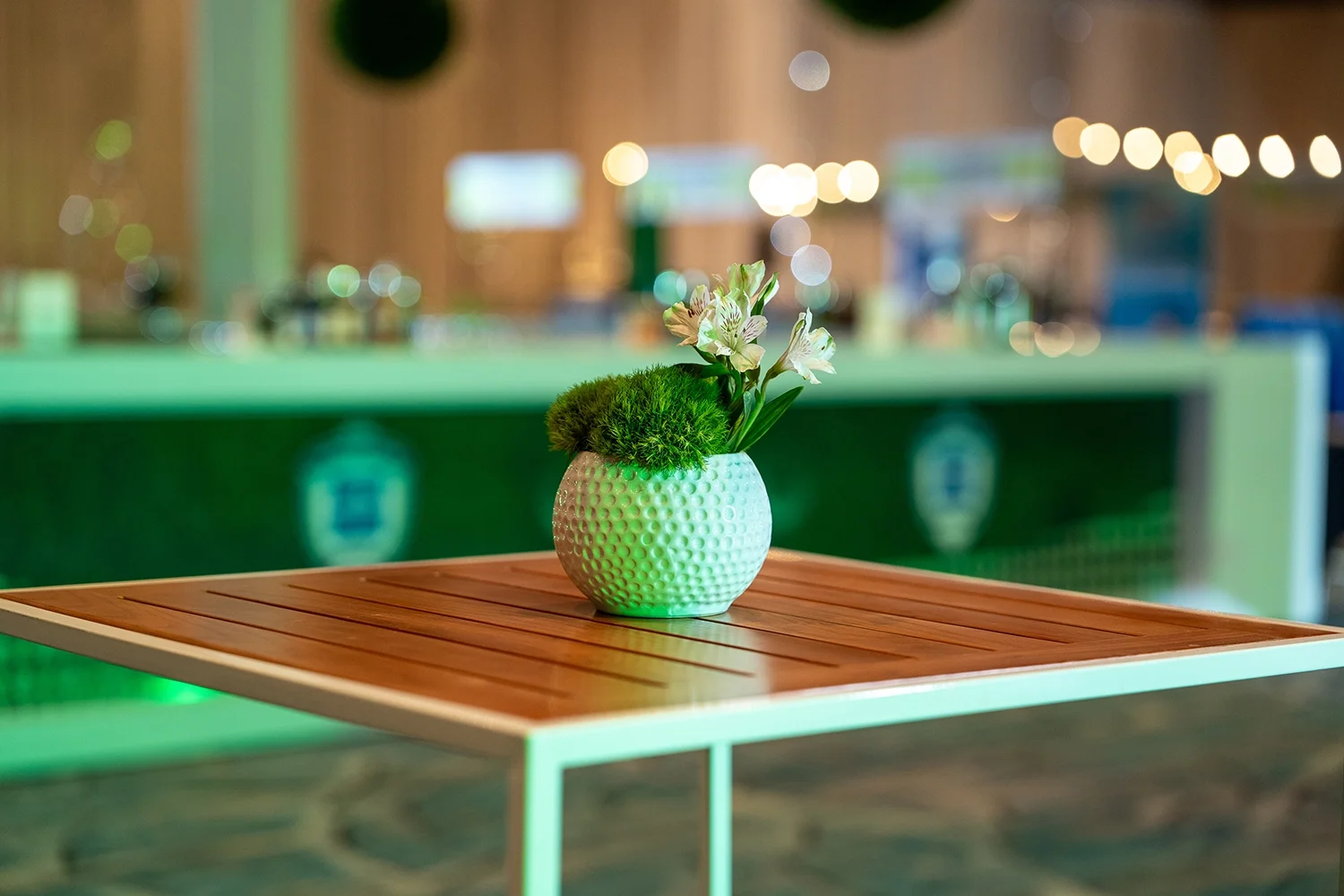 A white textured pot with green moss and white flowers inside, placed on a wooden table in an indoor setting with bokeh lights in the background.