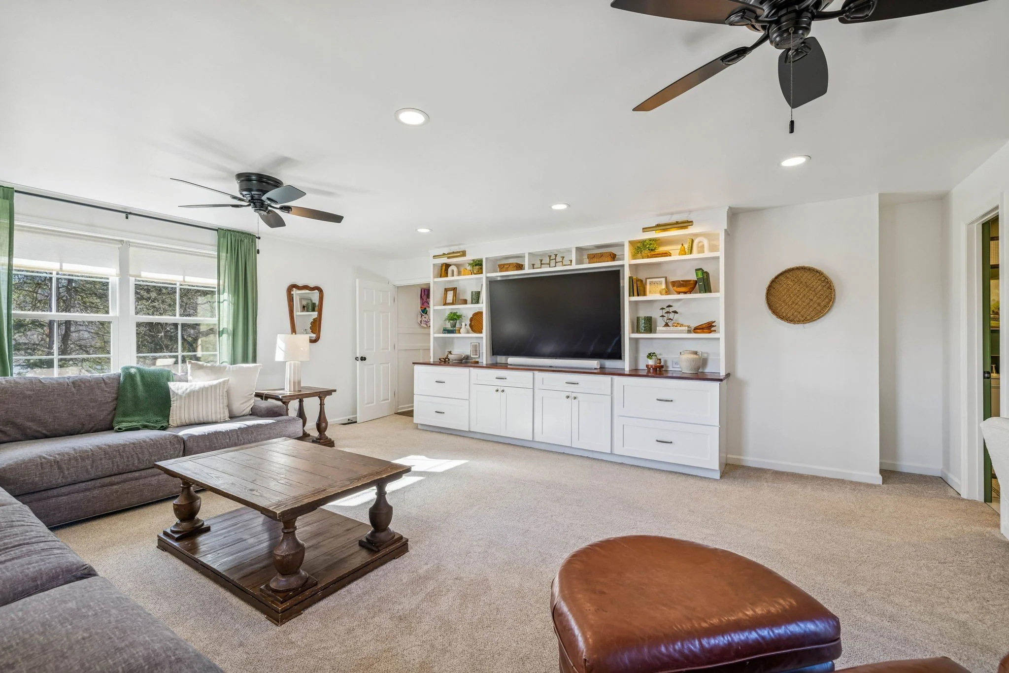 A living room with a large gray sofa, a wooden coffee table, and a leather ottoman. There are windows with green curtains, and a white built-in entertainment center with shelves, a television, and decorative items. Recessed lighting and two ceiling fans are on the ceiling.