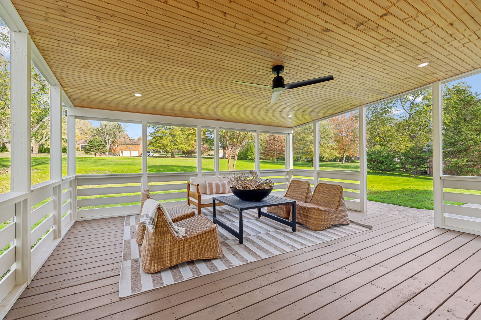 A screened porch with a wooden ceiling, ceiling fan, and outdoor furniture, overlooking a grassy yard with trees and houses in the background.