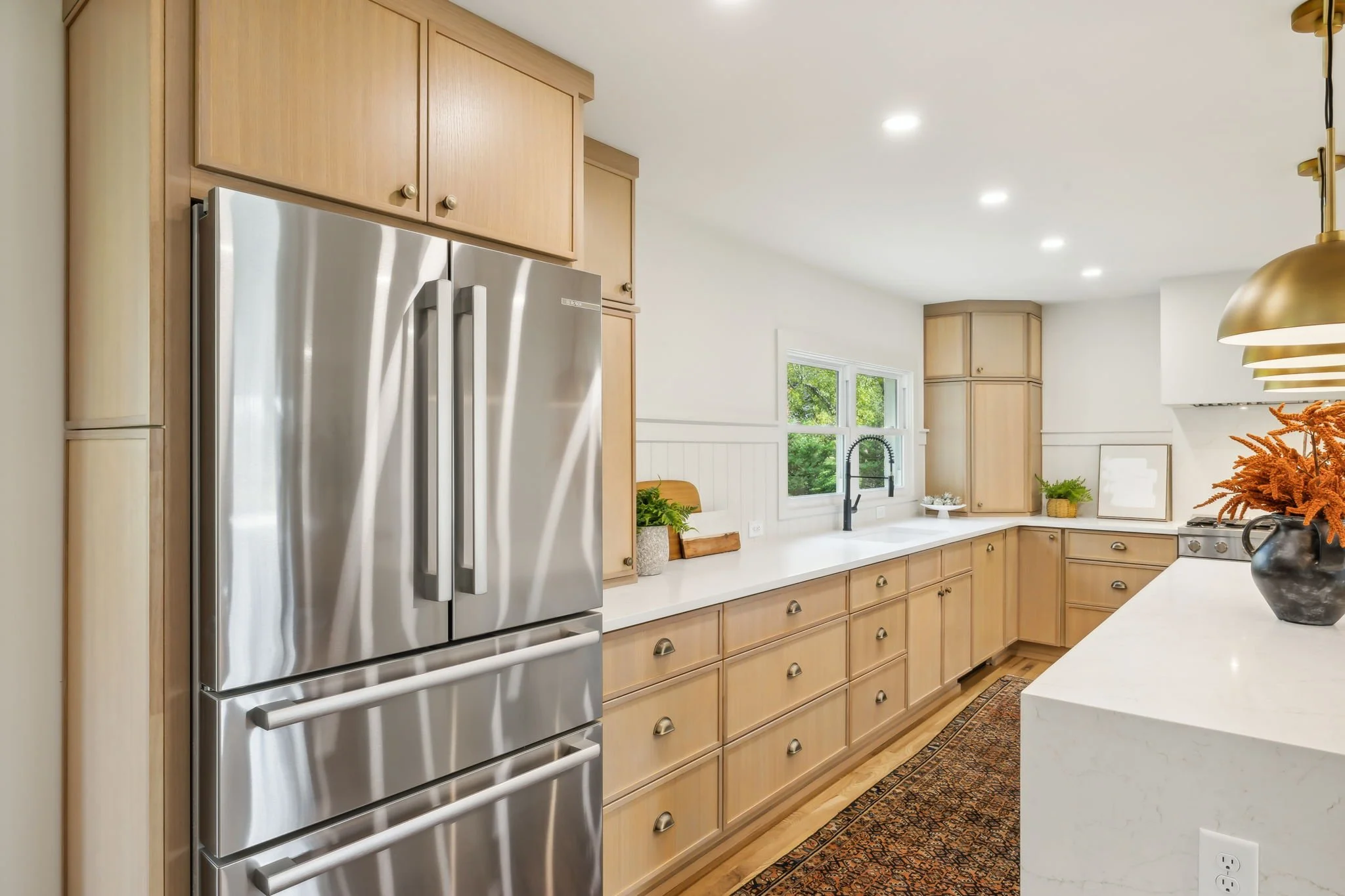 A modern kitchen with light wood cabinetry, a stainless steel refrigerator, white countertops, a window with greenery outside, and decorative items including a vase with orange flowers and a framed picture.