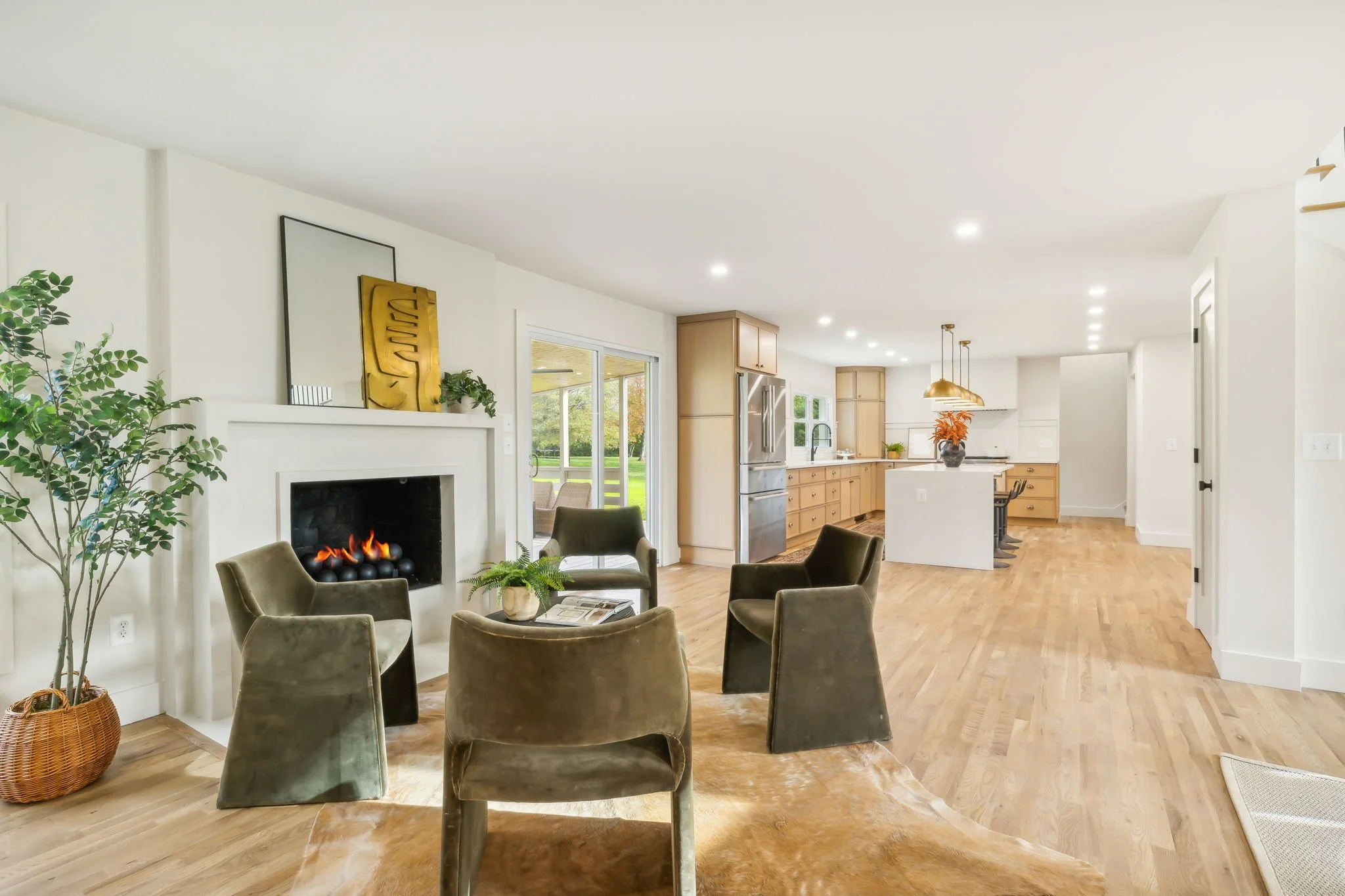Open-concept living room and kitchen with a fireplace, four green velvet chairs around a small table, plants, and large windows showing an outdoor view.