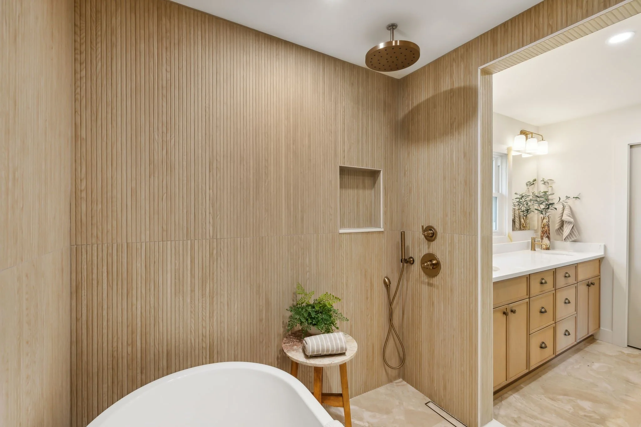Modern bathroom with wood-paneled wall, rainfall showerhead, brass fixtures, beige cabinetry, and a vanity with a mirror, potted plant, and towels.