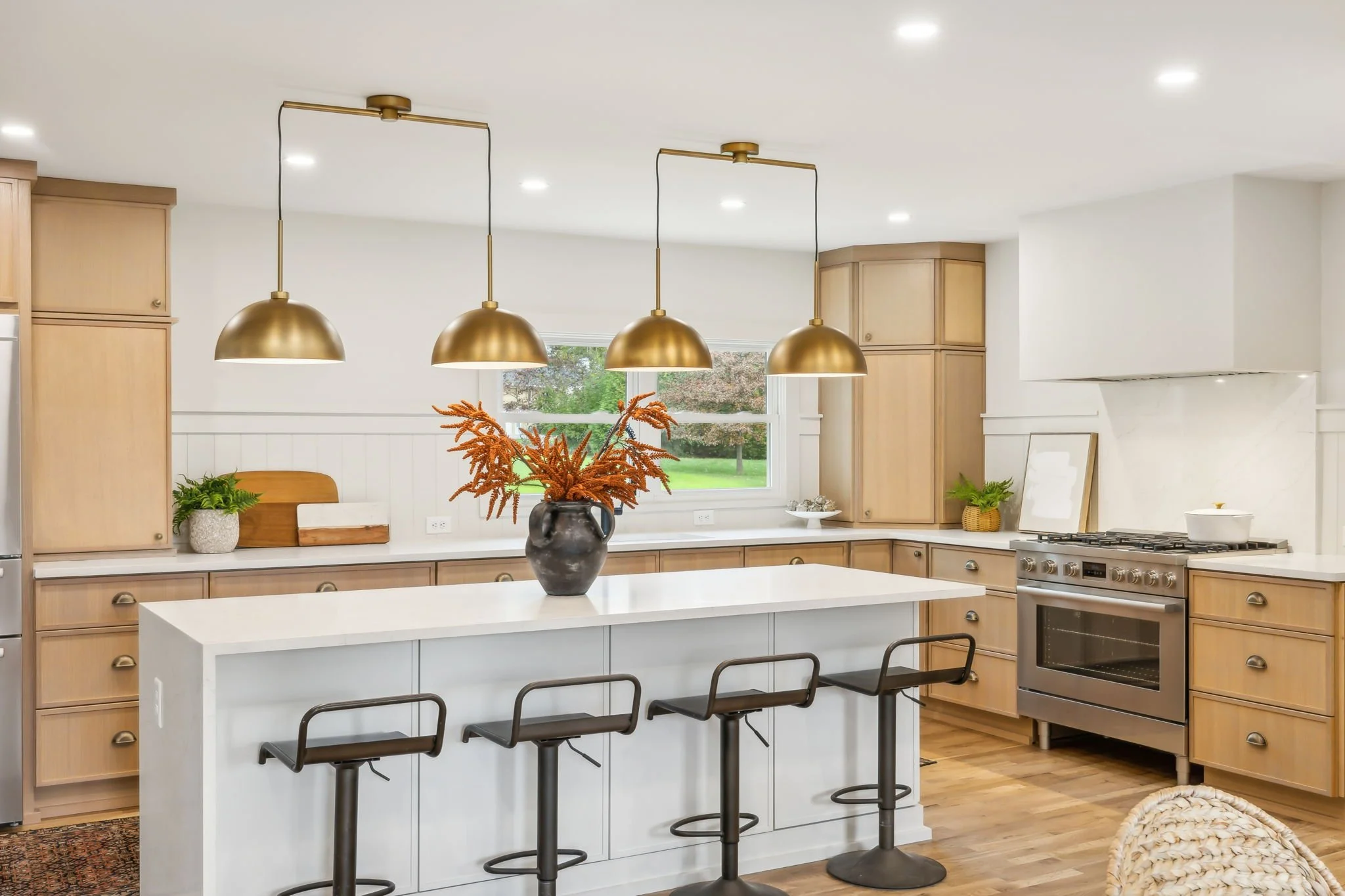 Modern kitchen with light wood cabinets, a white island with black bar stools, three gold pendant lights, and decorative plants.