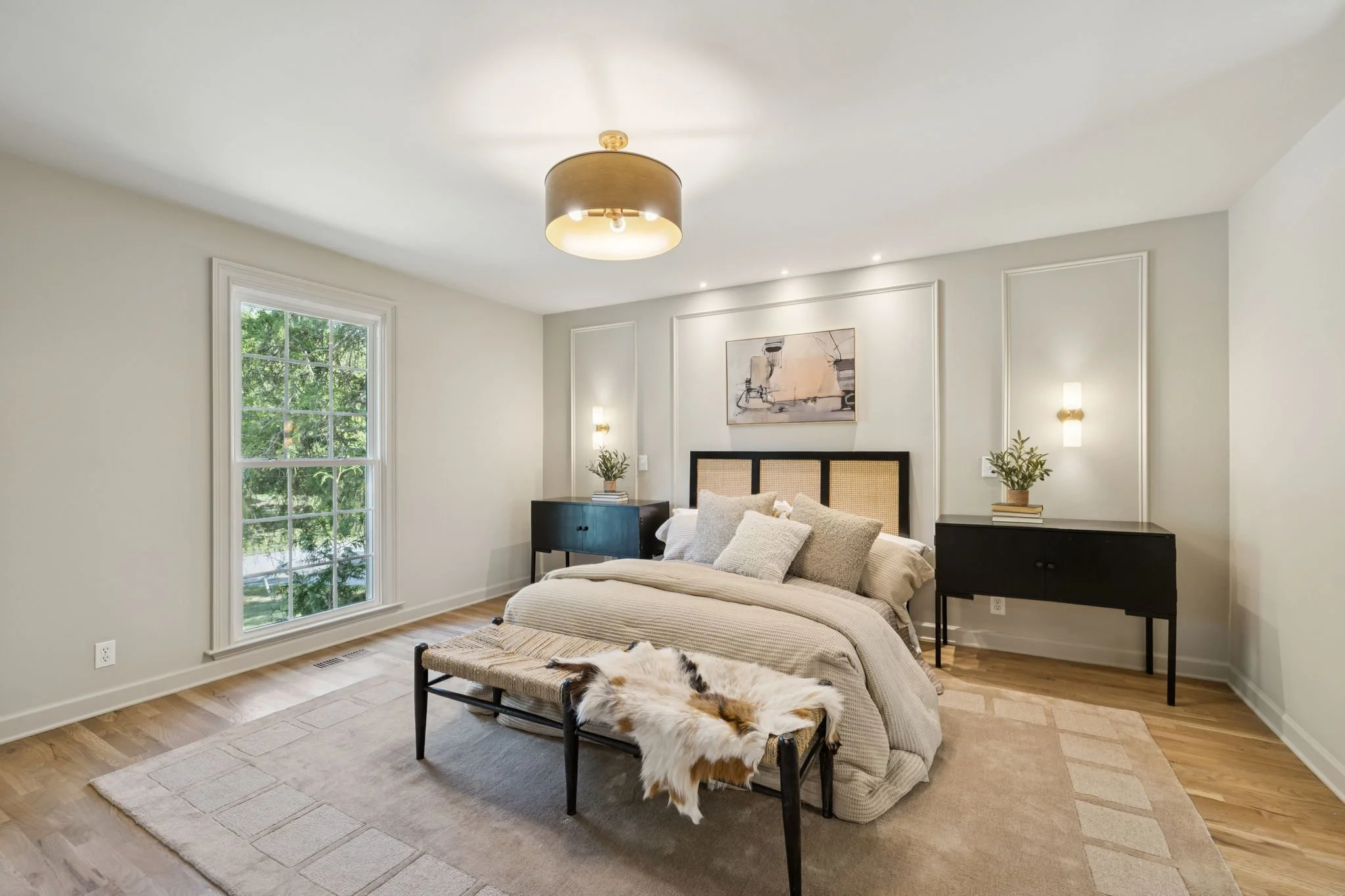 A bedroom with a large window on the left, a bed with beige bedding, a black side table, and paintings on the wall. There are plants on the side tables, and a fur throw on a stool at the foot of the bed. A cream-colored rug is on the wooden floor, with a modern light fixture on the ceiling.