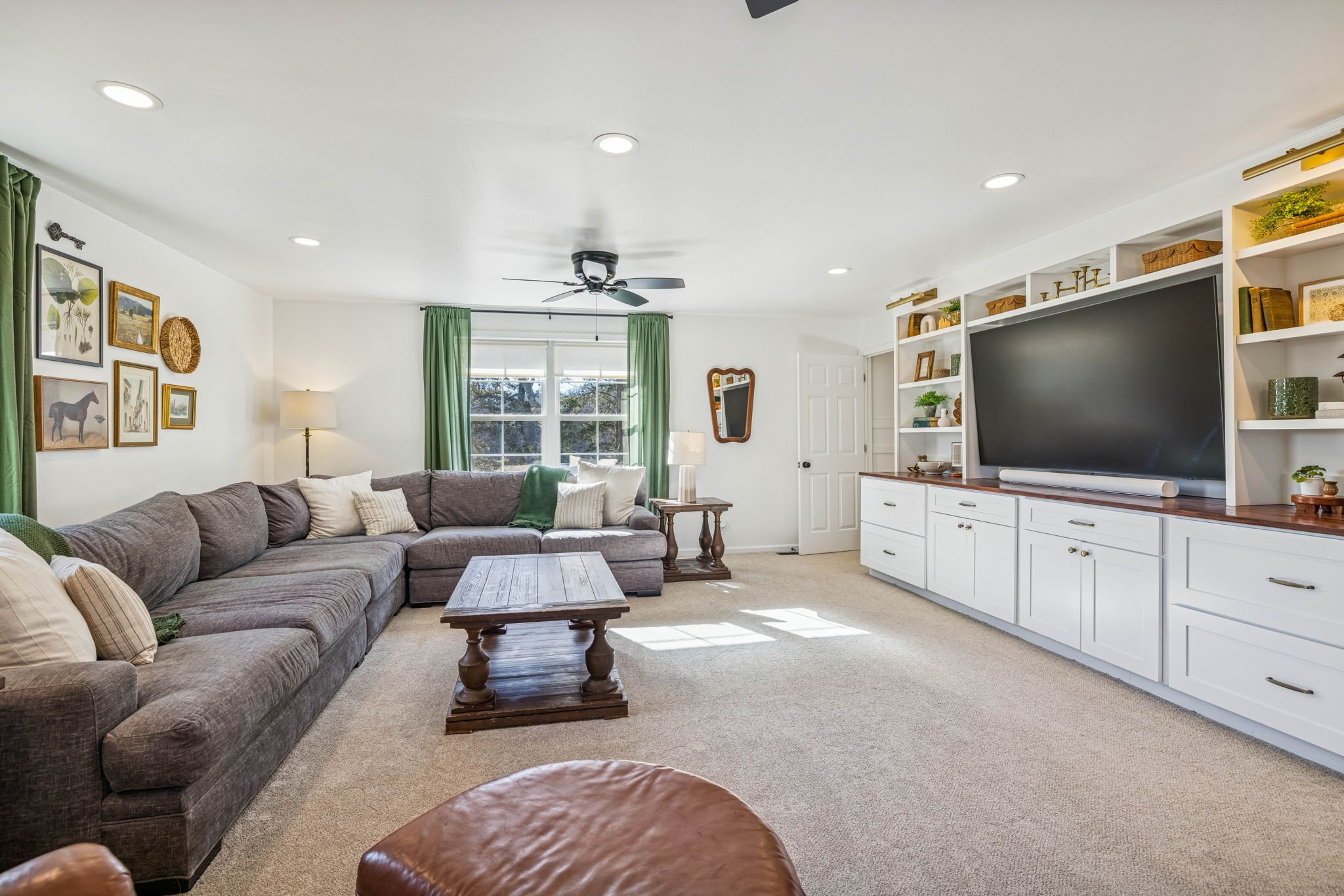 Living room with a large gray sectional sofa, a wooden coffee table, green curtains by a window, a TV on a white built-in unit with shelves, artwork and decorative items, a ceiling fan, and a beige carpet.
