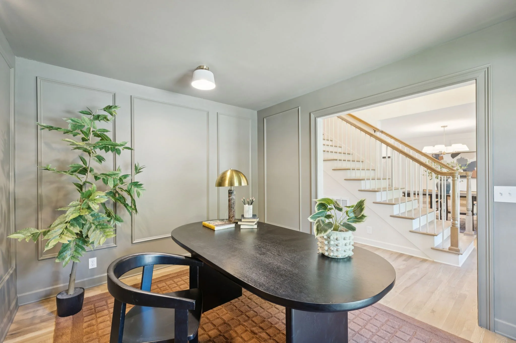 Interior view of a home dining area with a dark round table, surrounded by black chairs, decorated with plants, books, and a gold and white lamp. A staircase with wooden handrail and white balusters is visible through a large doorway. The room has light gray walls with decorative molding and a ceiling light fixture.