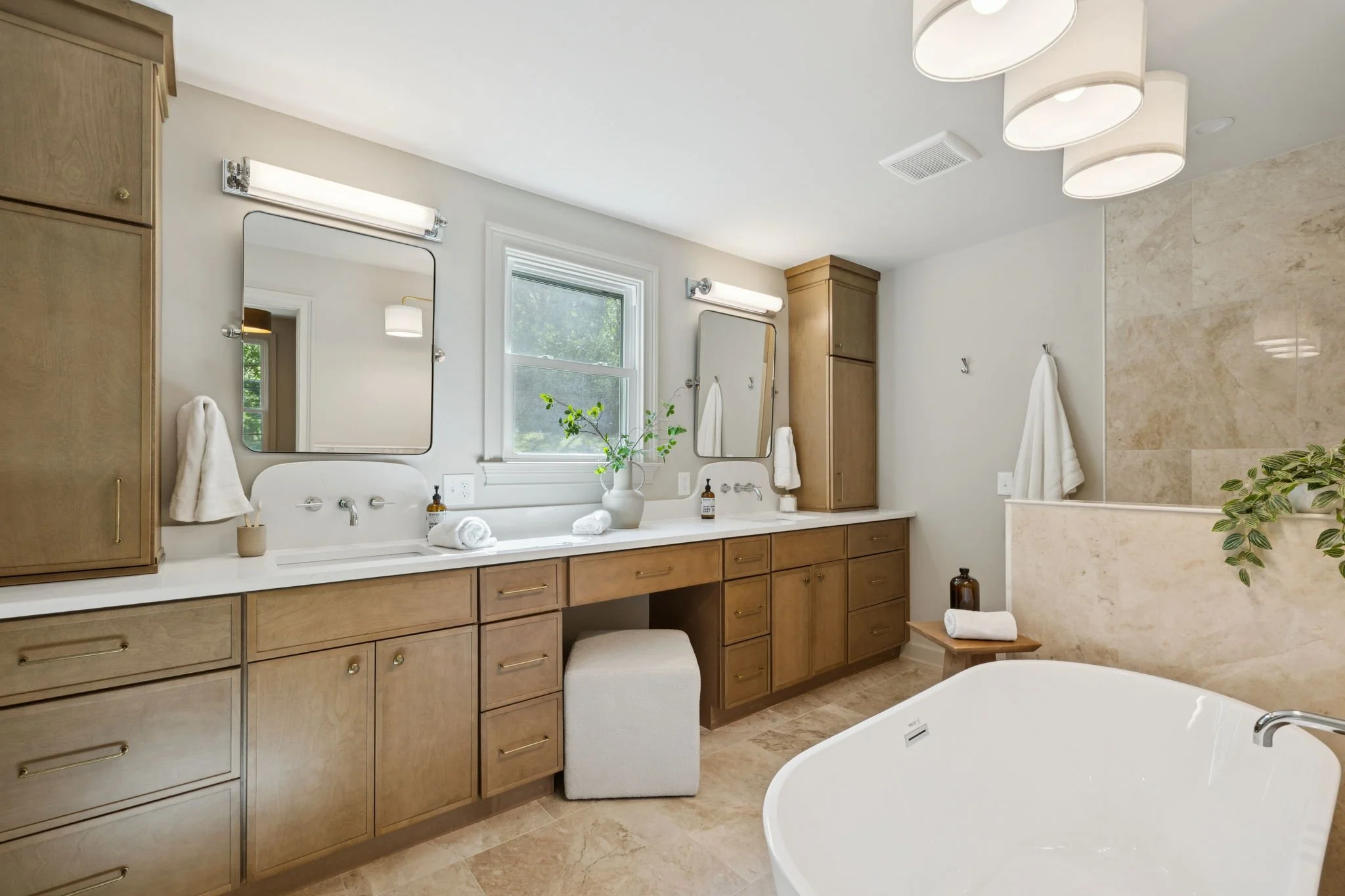 A spacious bathroom with wooden cabinets, a double vanity with two mirrors, wall-mounted lights, and a window with a plant on the sink. There is a bathtub on the right, and the room has a neutral color palette with beige and white tones.
