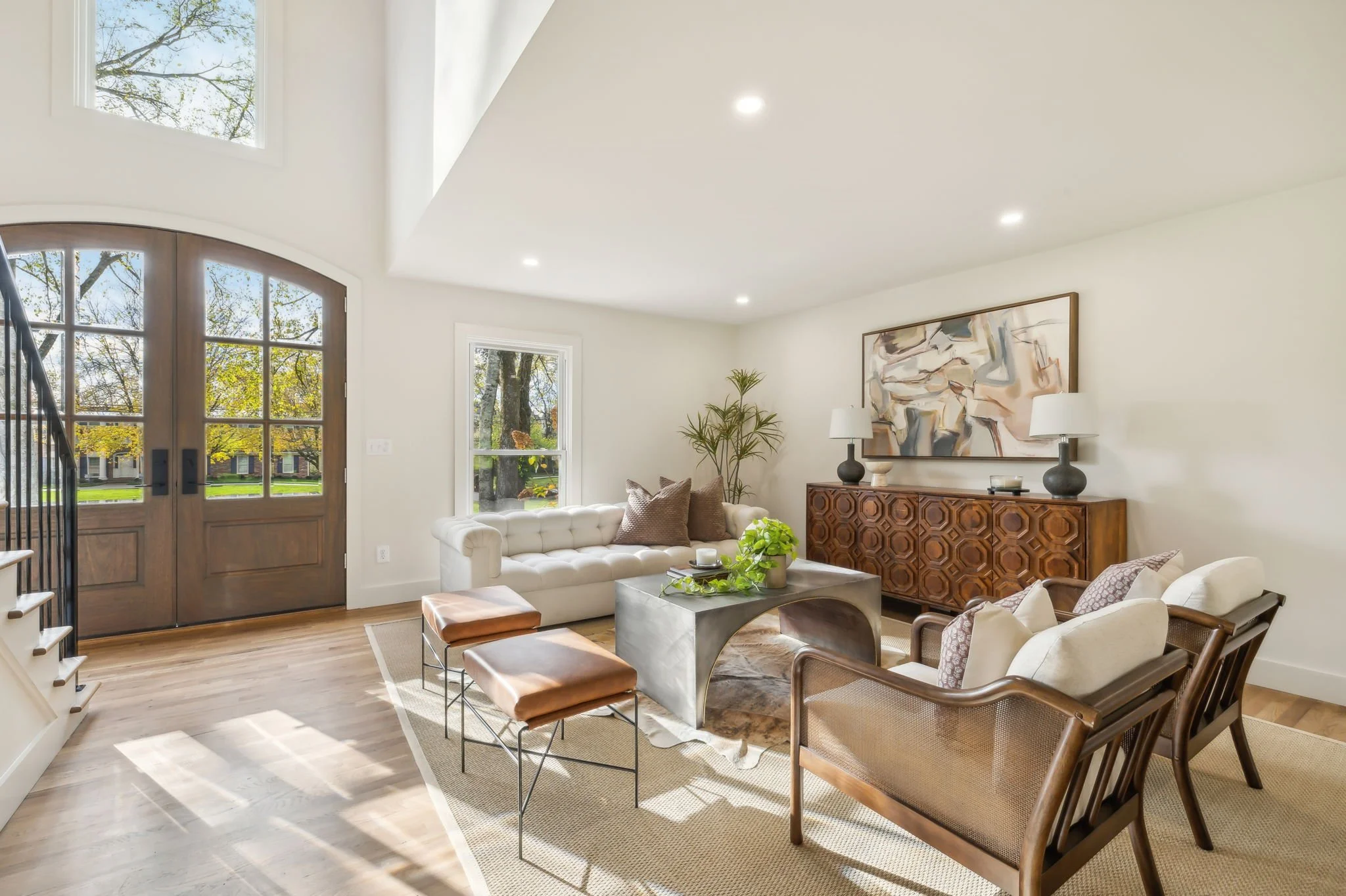 Living room with large wooden front door, white walls, hardwood floor, beige area rug, beige tufted sofa with throw pillows, glass coffee table with decorative items, two brown leather ottomans, side chairs, a large abstract painting, and large windows letting in natural light. Tree branches are visible through the windows.