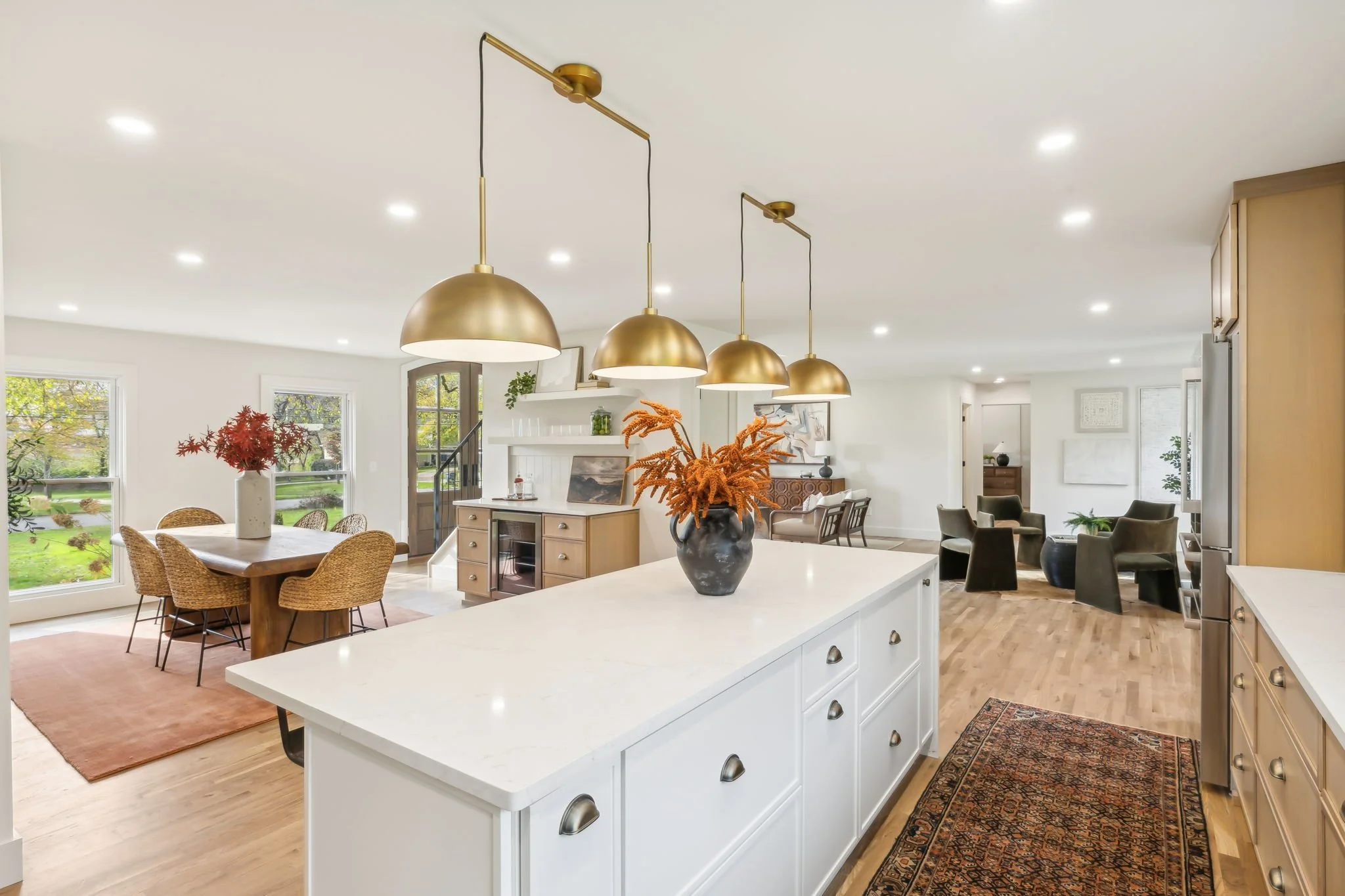 Open-concept living and dining room with white walls, hardwood floors, three metallic pendant lights, large windows with green trees outside, and various furniture including a dining table with wicker chairs, a kitchen island with decor, and a sitting area with armchairs and a side table.