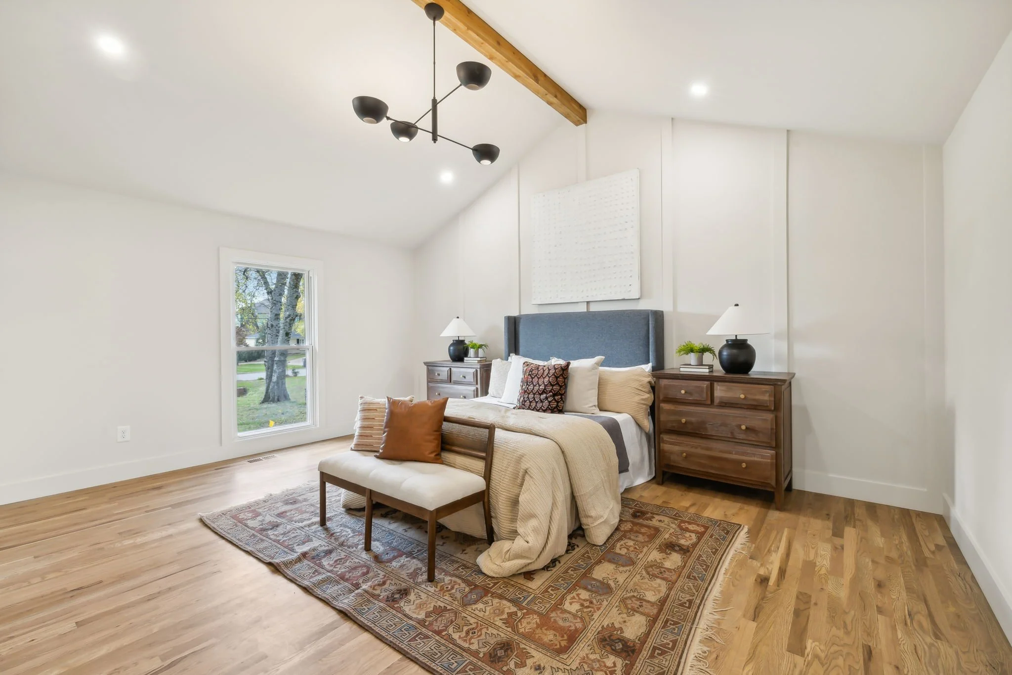 A bedroom with a wooden floor, a bed with a beige bedspread and multiple pillows, two wooden nightstands with lamps and plants, a window showing trees outside, and a modern black ceiling light fixture mounted on a wooden beam.