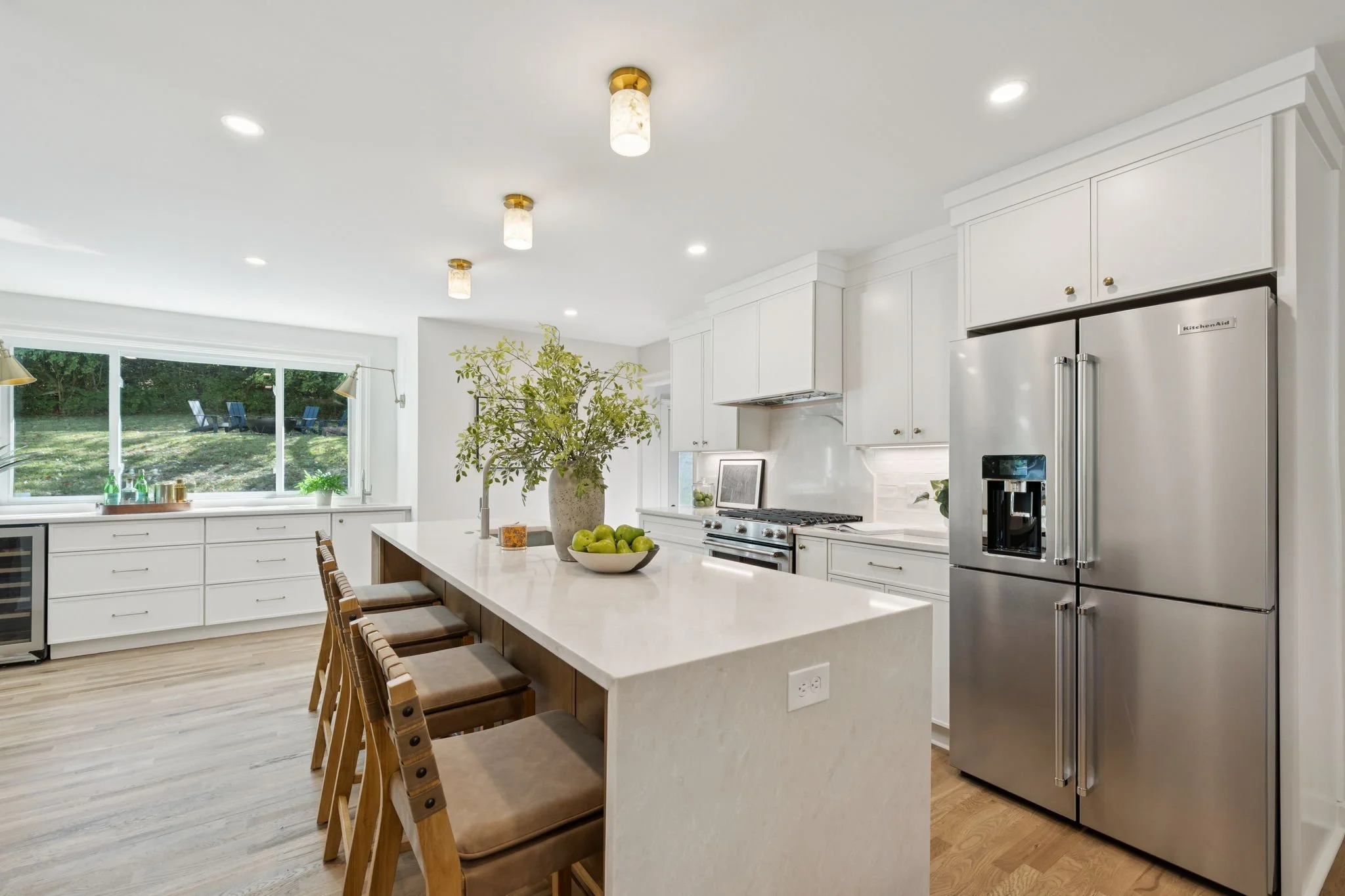 Modern white kitchen with large island, stainless steel refrigerator, and large window overlooking greenery outside.