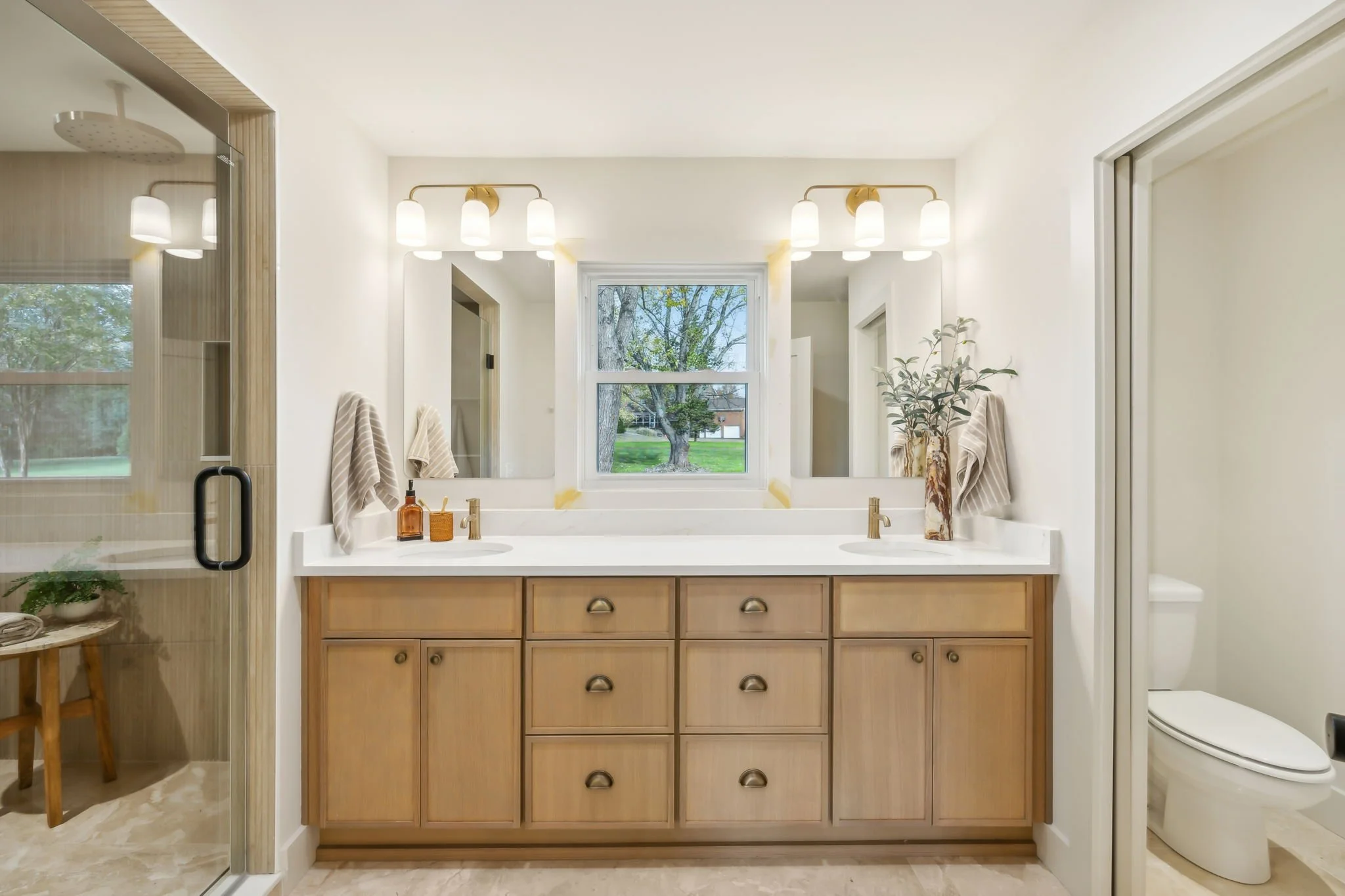 Bathroom with dual sinks, a large mirror, light fixtures, and a window showing trees outside. There are towels and decorative items on the counter.