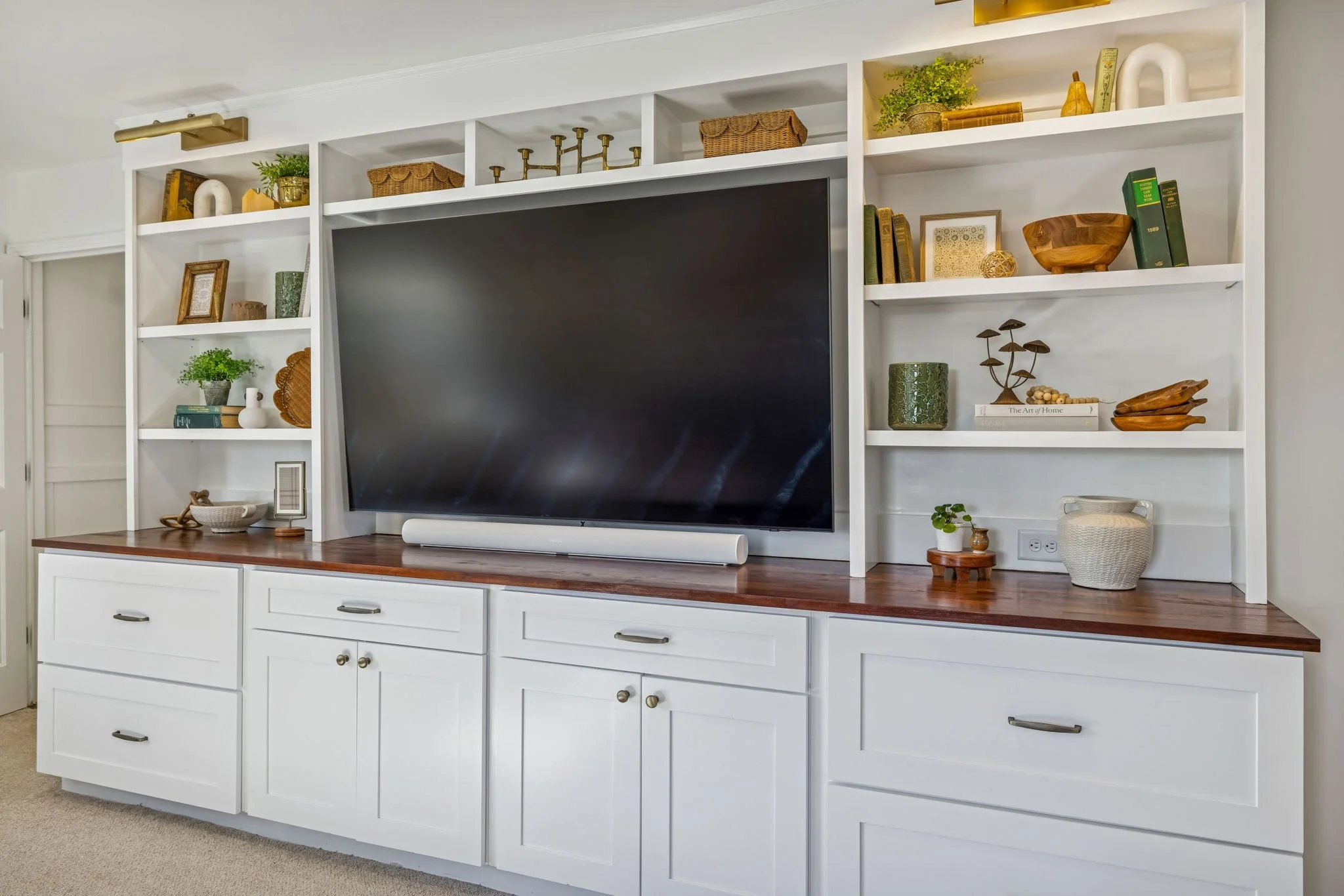 White built-in entertainment center with shelves filled with decorative items, books, plants, and a large flat-screen TV in the center.